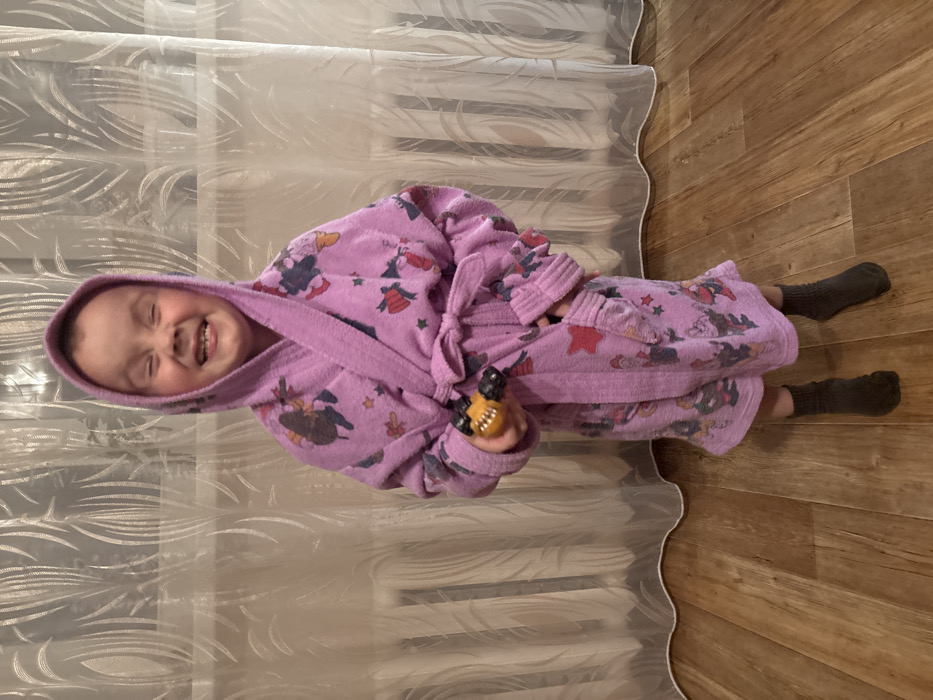 An indoor scene in Pavlovsky, Russia, depicts a young child standing centrally on a wooden floor. The child is wearing a purple hooded bathrobe with multicolored cartoon character and star patterns, tied at the waist. Their eyes are tightly squinted, and their mouth is open in a wide smile, revealing teeth. In their right hand, they hold a small yellow and black toy vehicle. The child wears dark-colored socks on their feet. The background consists of sheer white curtains featuring a vertical leaf-like pattern, behind which a white horizontal radiator is partially visible on the left side of the frame.