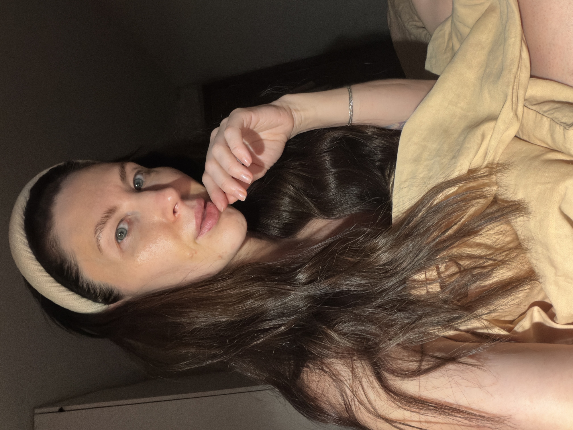 A woman with long brown hair and fair skin is lying down. She is wearing a beige headband and a beige shirt. She has a silver bracelet on her left wrist, and her left hand is touching her face. The background is dark, with a glimpse of a light-colored cabinet or wall in the lower-left corner. The location cannot be determined as Perm, Russia.
