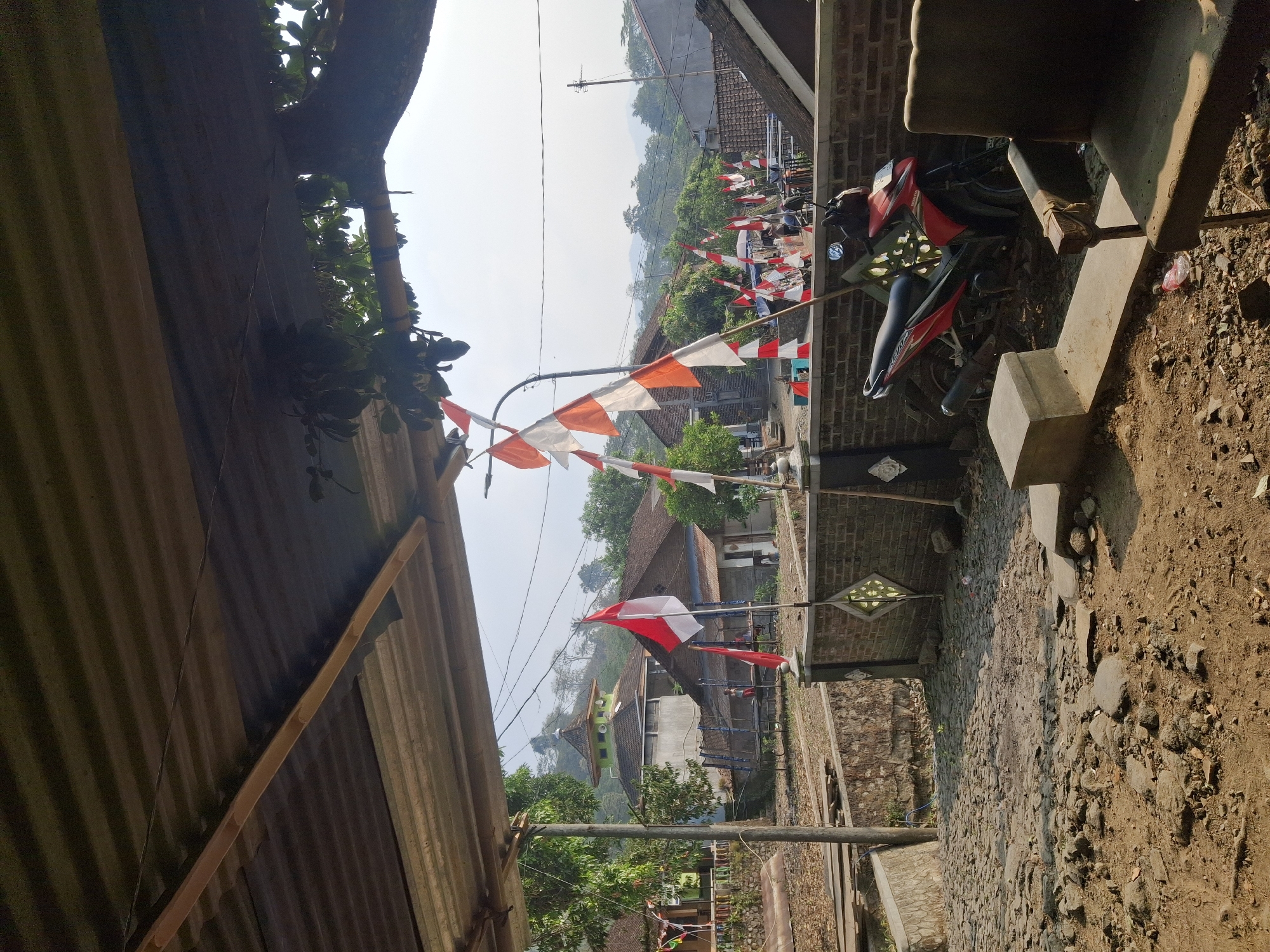 The image depicts an outdoor scene with a foreground dominated by rough, rocky ground and a dilapidated concrete structure. A red motorcycle is parked next to a brick wall adorned with a decorative diamond pattern. To the right, an aged chair with worn upholstery sits in the sunlight.
Beyond the immediate foreground, a cobblestone path leads towards a village setting. Several Indonesian flags, red and white, are prominently displayed on poles, suggesting a national celebration or commemoration. In the middle ground, a building with a green-tiled roof and a minaret-like structure stands out, possibly a mosque. Other buildings with traditional tiled roofs are visible, surrounded by lush green trees. The background reveals a hilly landscape under a hazy sky.
While the image provides a visual representation of a village environment with festive decorations, it does not contain explicit visual cues to pinpoint the exact location within the city of Muncar, Indonesia.