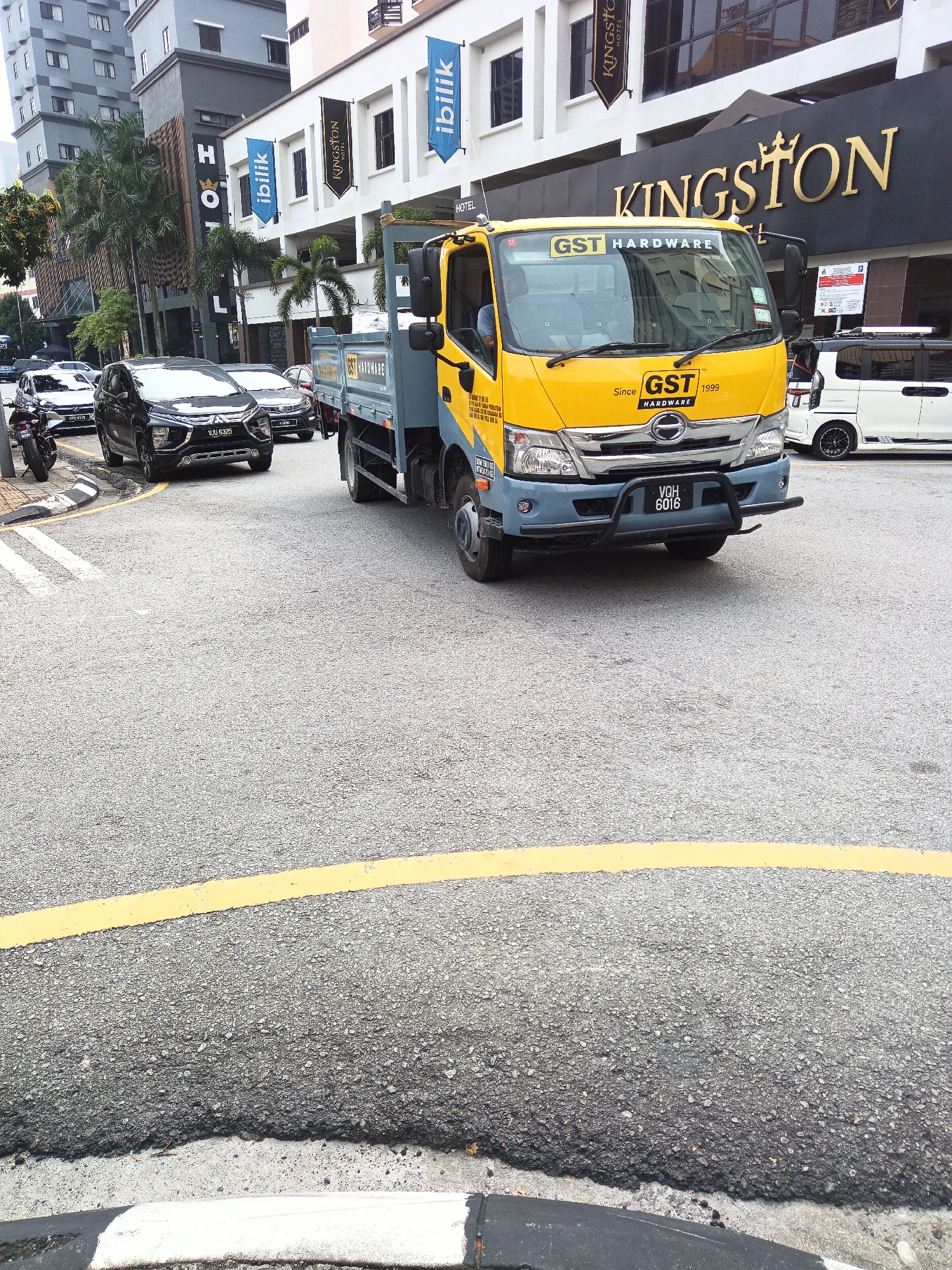 The image shows a street scene in Kuala Lumpur, Malaysia. A yellow and blue flatbed truck with the logo "GST HARDWARE" is the primary subject, parked on the right side of the road. The truck has the license plate VQH 6016. Behind the truck, a white commercial van with black accents is also visible. Several cars, including a black Mitsubishi Xpander with license plate VJU 6325, are parked or moving on the street. In the background, there are modern buildings, including a hotel with the "KINGSTON HOTEL" sign prominently displayed. Several flags and banners are visible on the buildings, including blue "ibilik" flags and a black "KINGSTON" banner. Palm trees are interspersed between the buildings, suggesting a tropical urban environment. The road surface is asphalt with yellow and white markings. The scene appears to be during daylight.
