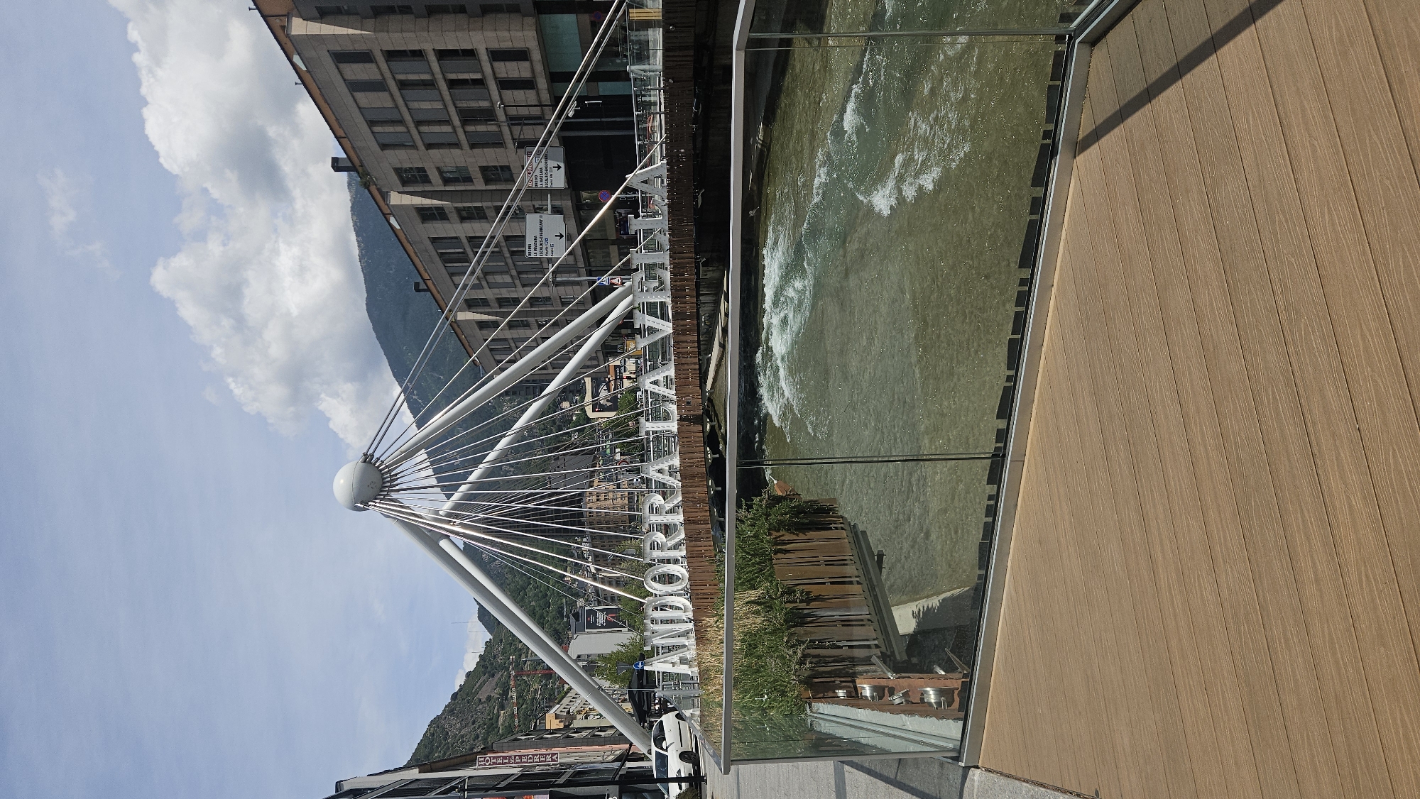 This image depicts the Pont de París (Paris Bridge) spanning the Gran Valira river in Andorra la Vella, Andorra, under a partially cloudy daytime sky. In the foreground, a light brown, wood-patterned composite deck extends diagonally from the bottom left, bordered by a clear glass barrier with a metallic frame that separates it from the river below. The Gran Valira river flows from right to left, exhibiting a greenish-brown hue with significant white rapids and ripples.
The midground is dominated by the pedestrian bridge, a modern cable-stayed design featuring a central, large white sphere from which numerous thin, metallic cables radiate downwards to support the bridge deck. Large, three-dimensional white letters spell "ANDORRA LA VELLA" across the side of the bridge deck, facing the viewer. Planters with green foliage are visible beneath the letters, both directly and reflected in the foreground glass barrier.
In the background, beyond the bridge, an urban landscape unfolds within a mountainous valley. To the left, older multi-story buildings with various architectural styles line a street, with a white car parked near the curb. One building prominently displays a red sign reading "HOTEL ANDREU." To the right, contemporary multi-story buildings with numerous rectangular windows and balconies rise. Steep, forested mountains form the distant backdrop, rising under a sky with both clear blue patches and scattered white cumulus clouds. Traffic signs are visible on the far side of the bridge, though their text is largely unreadable. No people are discernible in the scene.