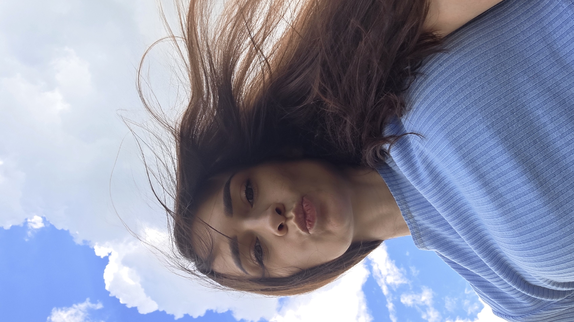 The image is a low-angle selfie of a young woman with long, dark brown hair blowing in the wind, set against a bright blue sky with scattered white clouds. The woman is looking directly at the camera with her lips pursed as if blowing a kiss. She is wearing a light blue ribbed top. The overall scene suggests an outdoor setting, possibly in a park or open area, typical of the village of Daryino, Russia.