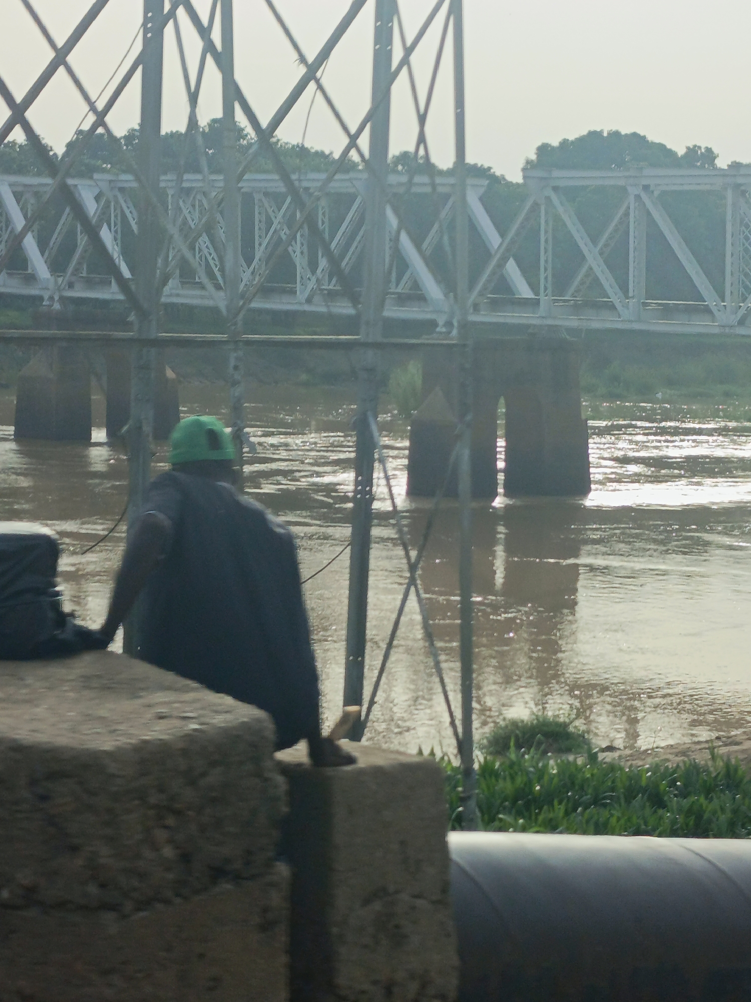 The image captures an outdoor scene in Barnawa, Nigeria, featuring a person situated on a concrete structure, observing a large metal truss bridge spanning a wide river. The individual is viewed from behind, wearing a green cap and a dark garment, and is positioned on the left side of the frame, facing towards the right. Multiple vertical metal posts with cross-bracing stand between the foreground and the main bridge. The background is dominated by a light-colored, multi-span truss bridge constructed of metal, supported by dark, thick pillars extending into the water. The river exhibits brown, moving water with numerous reflective highlights. Green trees are visible on the distant riverbank behind the bridge, while lower green vegetation lines the closer bank. A large, dark, curved pipe is partially visible in the lower right corner. The lighting suggests an overcast or hazy sky.