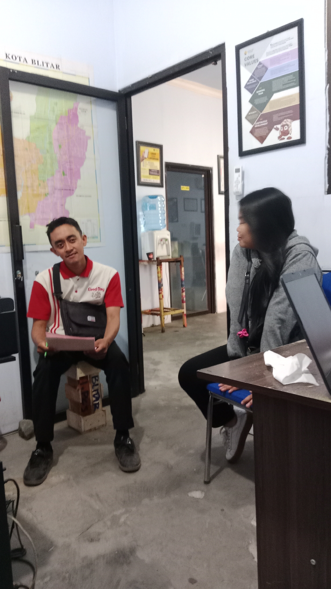 The image depicts an indoor scene, likely an office or meeting room, in Blitar, Indonesia. A man in a red and white collared shirt and black pants is seated on a stack of wooden crates, holding a pink notepad and a pen. He has a black messenger bag slung across his chest. To his right, a woman with long dark hair, wearing a grey hooded jacket and black pants, is seated in a blue chair at a dark wooden table. A white tissue is on the table in front of her. In the background, a map of Blitar city is visible on a wall, along with framed posters and a water dispenser. The environment appears to be a functional workspace with concrete flooring.