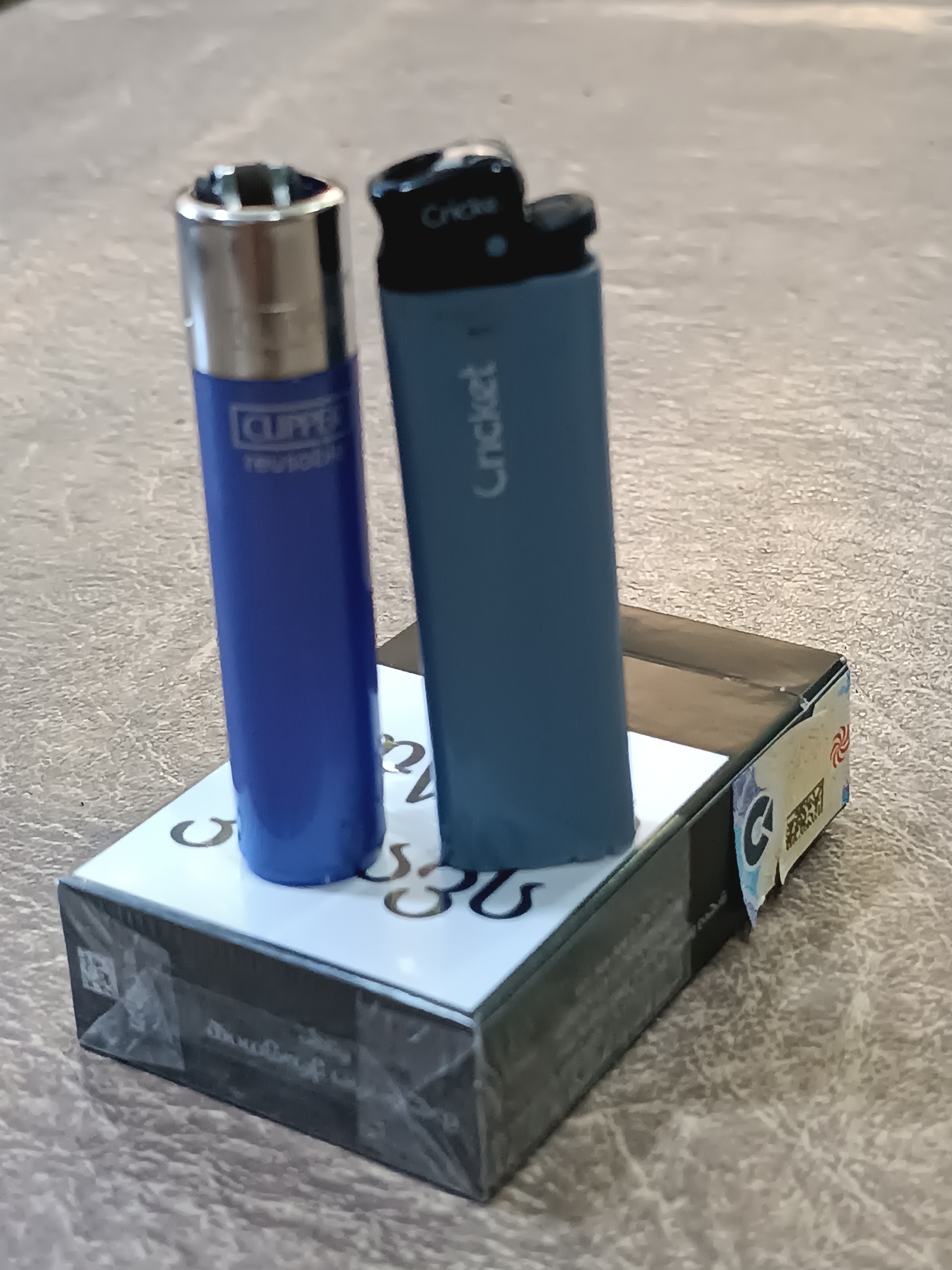 The image shows two cigarette lighters, one blue and one teal, resting on a pack of cigarettes. The blue lighter is a Clipper brand, and the teal lighter is a Cricket brand. The cigarette pack is black and white with some Georgian text and graphics. The lighters and cigarettes are placed on a textured surface, possibly a table or countertop. There are no people visible in the image. The scene is indoors, and the lighting appears to be natural. The image does not provide any specific information about the city of Khashuri, Georgia.