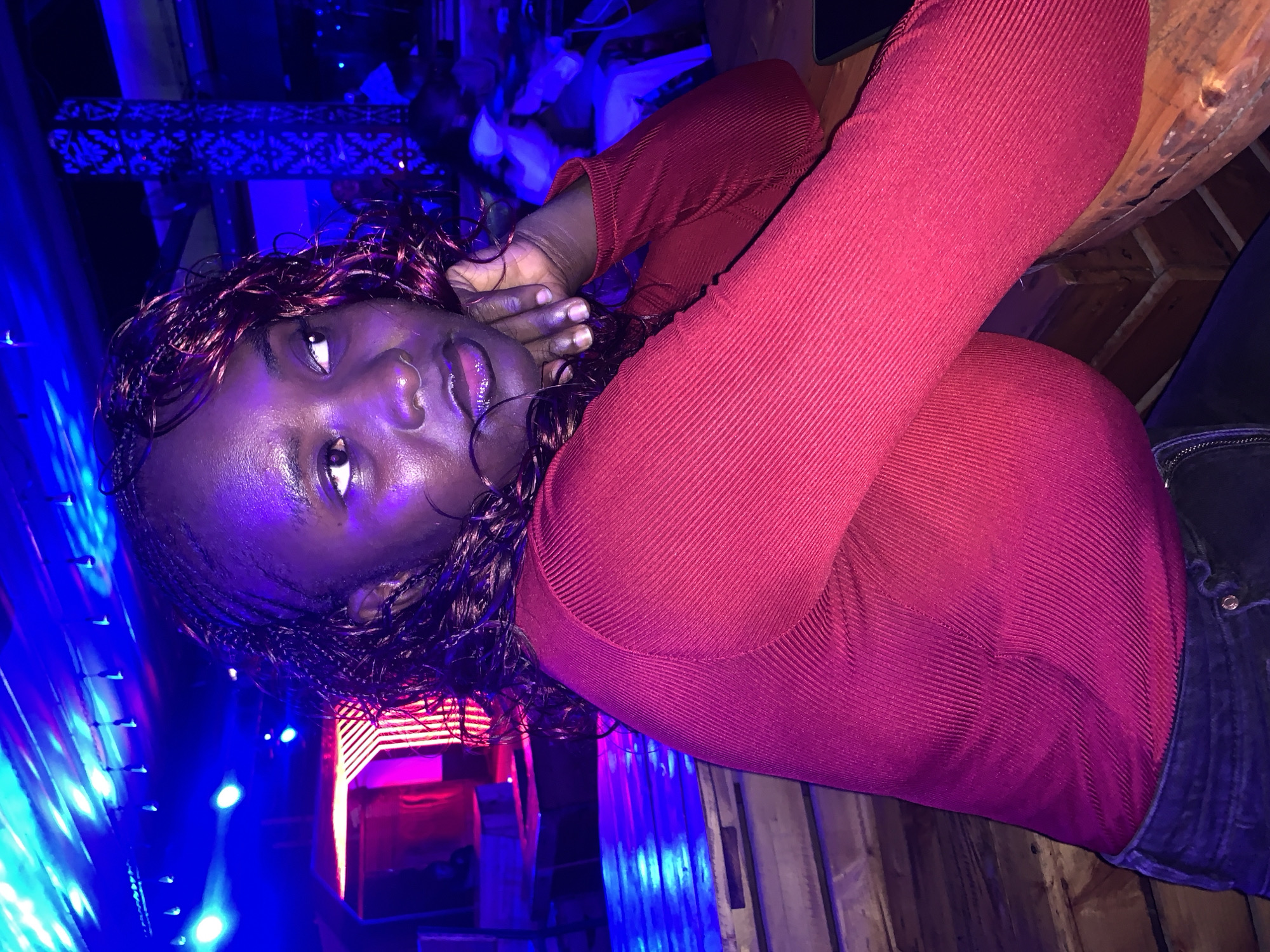 A young Black woman with braided hair and deep red colored streaks sits at a wooden table in a dimly lit establishment. The scene is bathed in vibrant blue and pink lighting from overhead fixtures and a neon sign in the background. She is wearing a long-sleeved, ribbed, deep red top and dark jeans. Her chin rests on her clasped hands, and she is looking to her right, her gaze directed slightly upwards. Her lips are glossy, and her skin has a dewy sheen under the colored lights. In the background, blurred figures and architectural elements are visible, including patterned columns and what appears to be a stage or elevated area with glowing pink lights. The overall ambiance suggests a nightclub or bar. The specific location in Howayi, Nigeria, cannot be determined from the visual information provided.