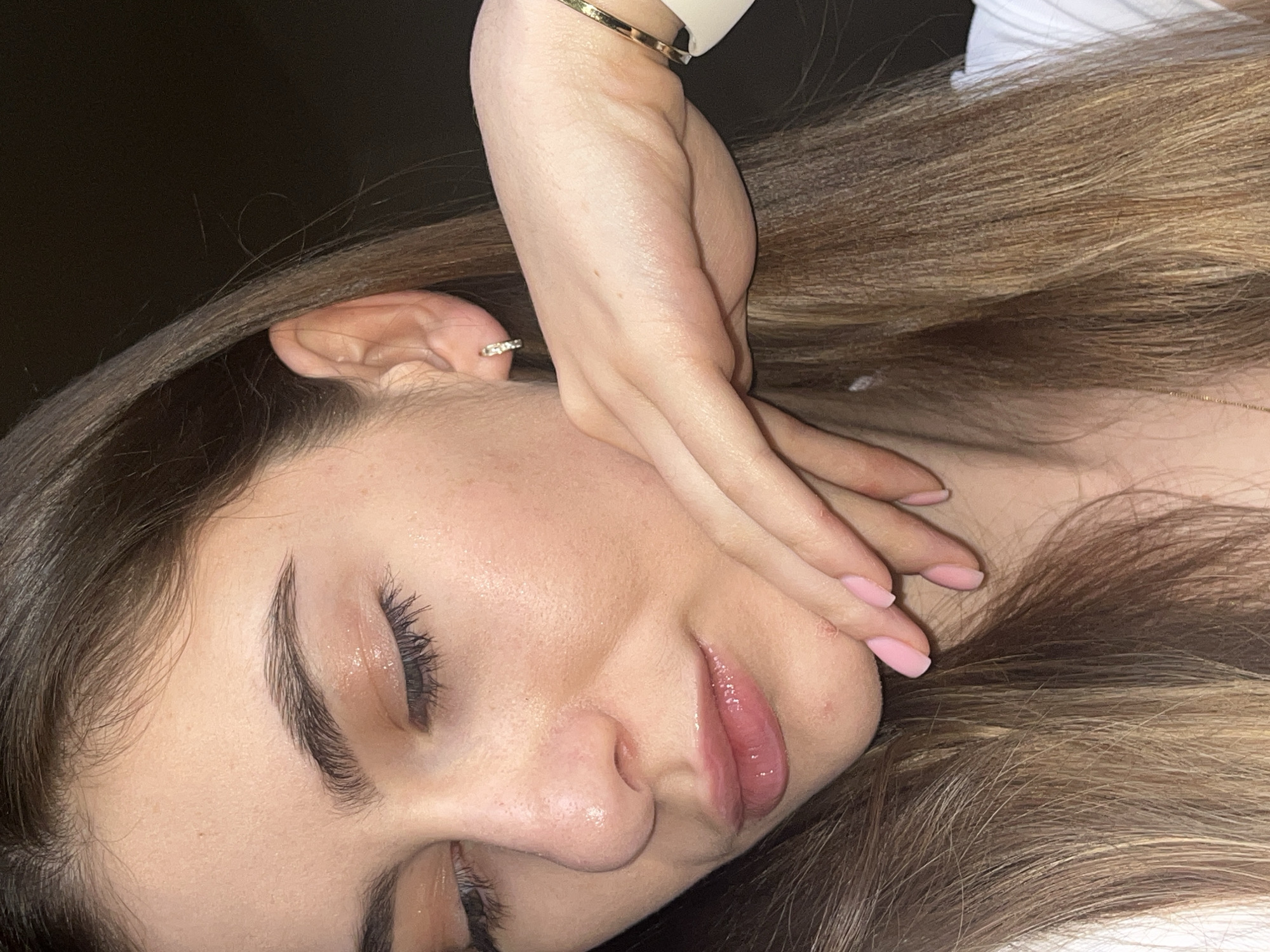 A close-up photograph depicts a person with light brown hair. The individual's left ear is visible, adorned with a circular earring. The person's left hand is placed near their face, with pale pink fingernails. The image appears to be taken indoors, with a dark background in the upper-left corner. The setting is in the city of Unknown, Unknown.