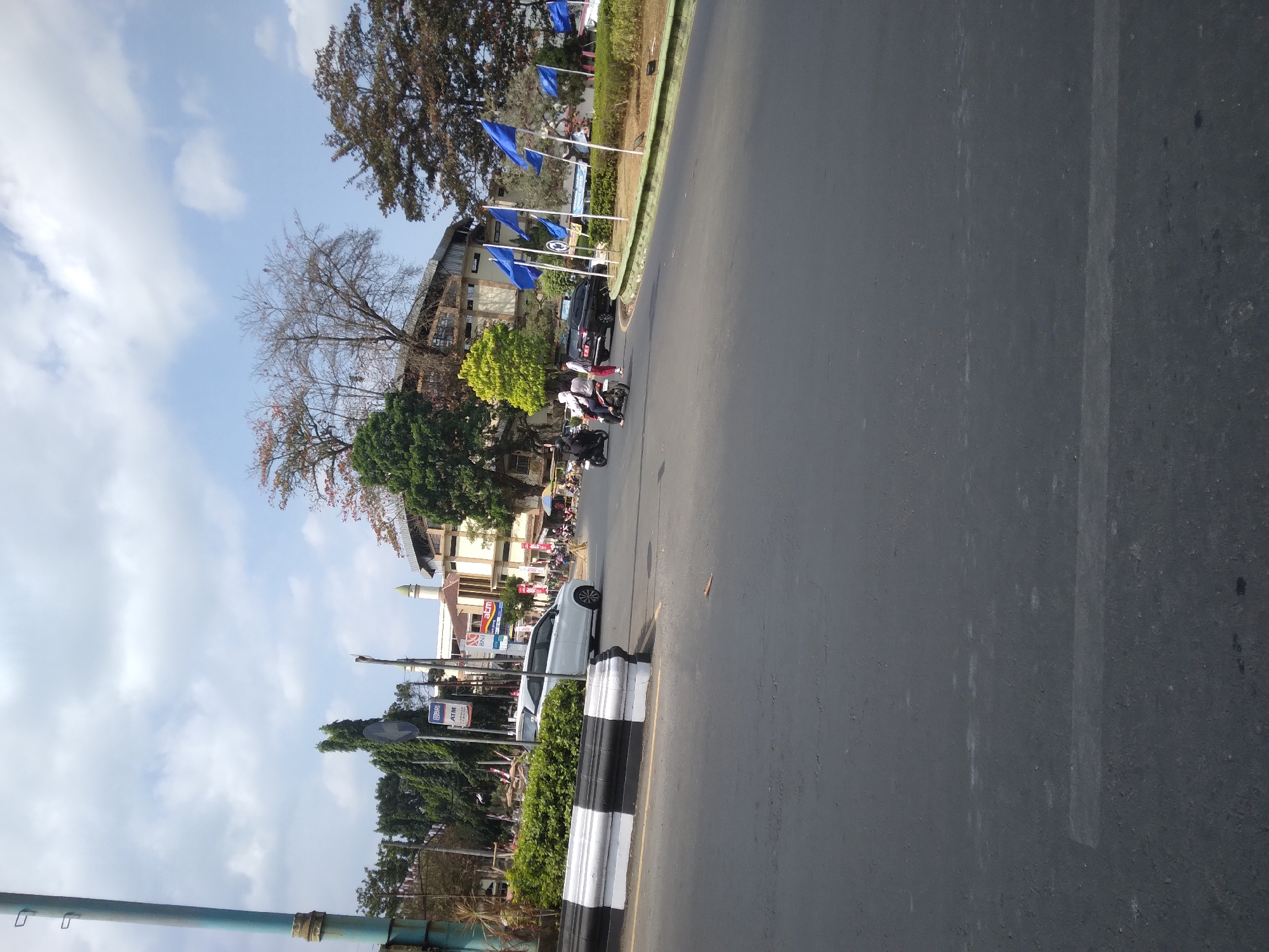 The image shows a street scene in Sokaraja, Indonesia, with a clear sky and scattered clouds. In the foreground, a paved road is visible with lane markings. On the left side of the road, there is a black and white striped barrier and a median with green bushes. A tall blue pole stands on the far left.
Further down the road, several vehicles are present, including a white car and a black car. Multiple motorcycles are also seen, with riders on them. A few people are walking or standing near the vehicles. In the background, there are buildings, including what appears to be a mosque with a green dome. Trees of various sizes and types are scattered throughout the scene. Several blue flags on poles are lined up along the median. The overall impression is of a moderately busy street in a commercial or residential area.
