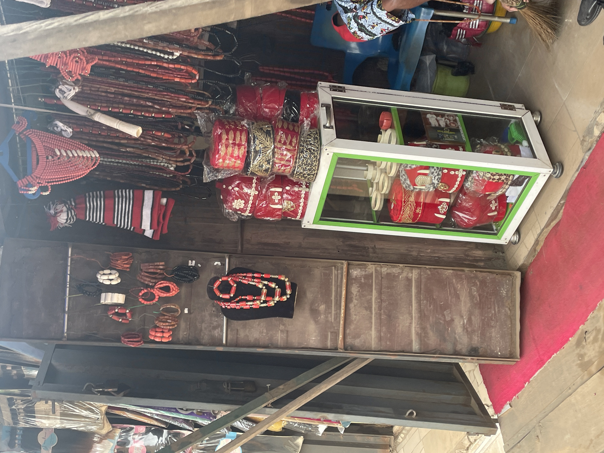This image captures a market stall in Nkpor, Nigeria, showcasing a vibrant display of traditional Nigerian accessories. The stall, constructed from wood and metal, features a collection of red beaded necklaces and bracelets, arranged on a mannequin and hanging from racks. Adjacent to the jewelry, stacks of red and black hats, adorned with gold embroidery and shells, are displayed in a glass-fronted cabinet. In the background, a person dressed in colorful traditional attire can be seen sitting on a blue plastic chair. The ground is covered with a red carpet, and a broom is visible in the foreground, suggesting ongoing activity in the market. The overall scene portrays a bustling commercial environment characteristic of Nigerian markets.