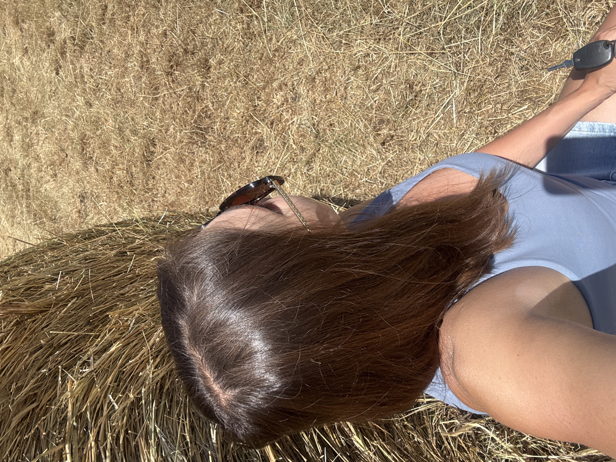 The media file shows a person lying on a haystack in an outdoor setting. The subject is wearing a light blue top and denim shorts. Sunglasses are perched on their head. A set of car keys rests on their thigh. The person's brown hair is spread across the haystack. The sun is shining brightly, casting shadows and highlighting the texture of the hay. The scene suggests a warm, sunny day, possibly in the countryside. The orientation of the image is from a high-angle, looking down at the person. The overall mood is relaxed and casual. There is no visible text in the media file.
