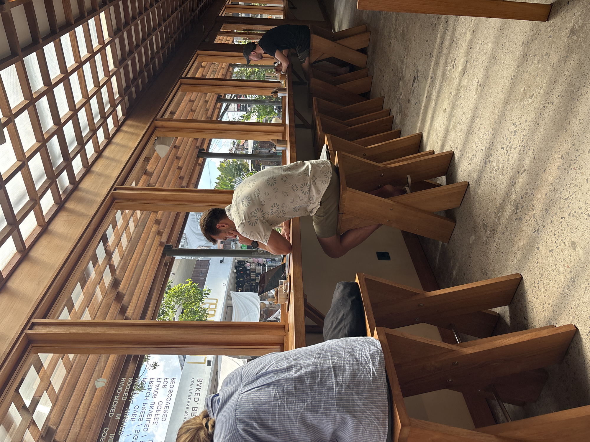 The image shows a group of people sitting at a long wooden counter in a cafe. The cafe has a wooden-framed glass wall, allowing natural light to fill the space. The people are sitting on unique wooden stools that appear to be formed by angled wooden beams. One man is wearing a light-colored shirt with a pattern and tan shorts. Another person, mostly out of view, is wearing a blue striped shirt. A third person is wearing a black shirt. The cafe appears to be "Baked," located in North Kuta, Indonesia, based on the reflection in the window.