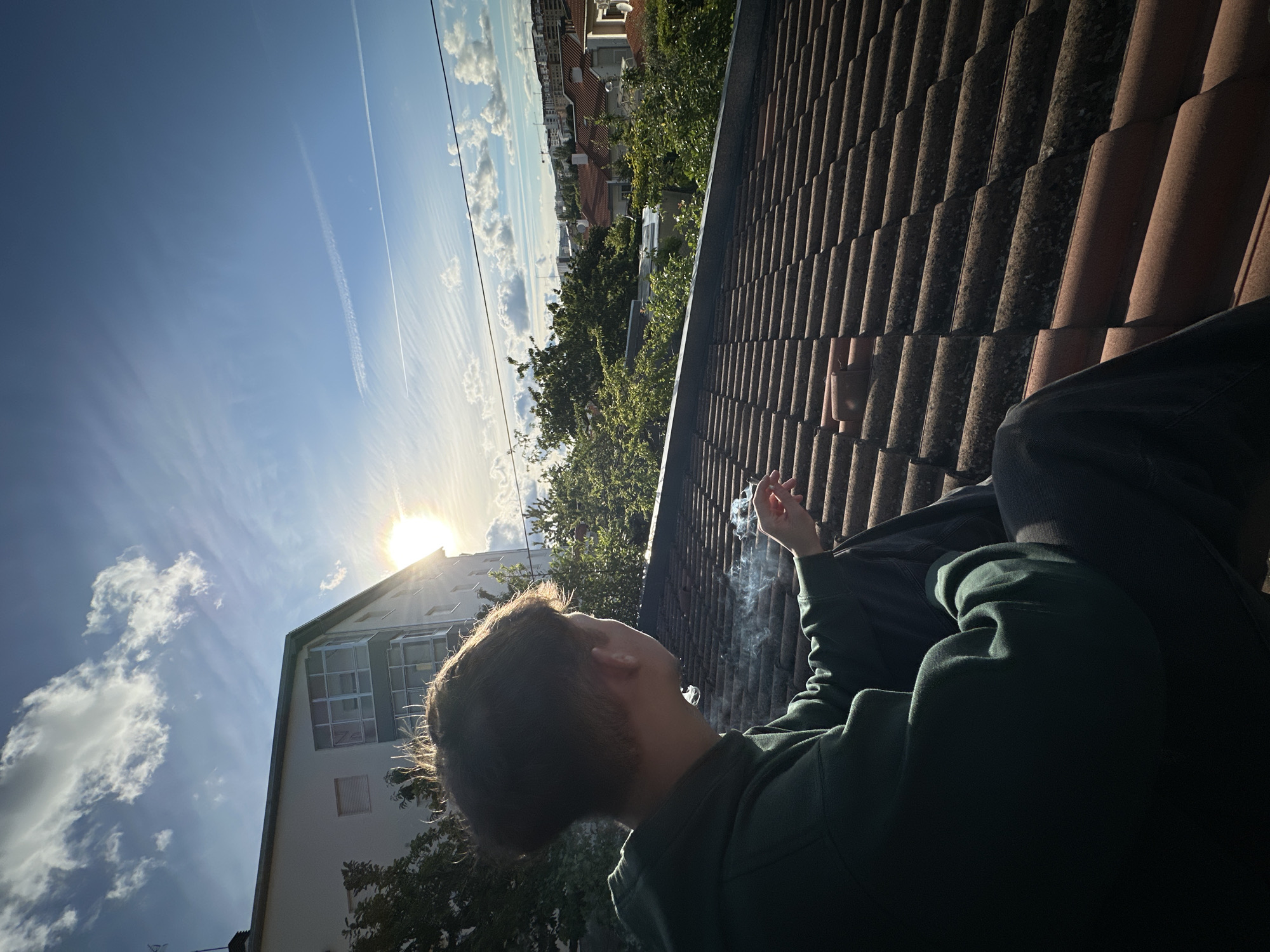 The image is taken from a low angle, looking upwards towards the sky. In the foreground, a person with short brown hair, wearing a dark green hooded sweatshirt and dark pants, is seated on a tiled roof. The person is holding a lit cigarette in their right hand, and smoke is visible emanating from the cigarette.
The background consists of a partly cloudy blue sky with contrails from an aircraft, a building with white exterior walls and several windows, and green trees that surround the buildings. In the distance, other buildings can be seen.
While I cannot verify the location is in the city of Bragança, Portugal, there is nothing in the visual content to contradict this.