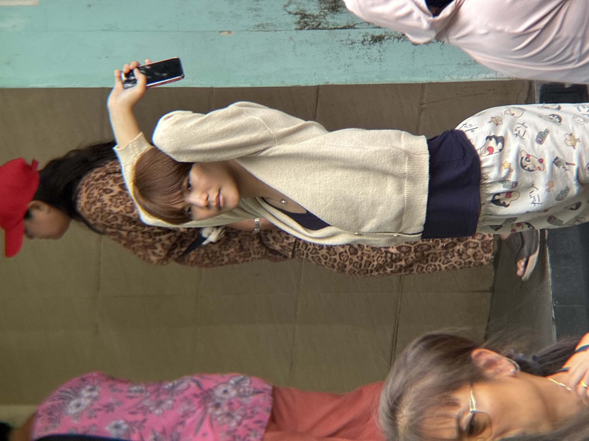 In Lao Bảo Commune, Vietnam, a young woman with short, dark hair and a fair complexion is captured mid-action, holding a black smartphone in her outstretched right hand. She is wearing a cream-colored cardigan over a dark navy top, and white patterned pants featuring cartoon characters. The woman is standing in an outdoor urban environment, with a textured, light brown wall behind her and a teal-colored wall to her right. Two other individuals are partially visible in the background: one to the left wearing a bright pink patterned shirt and an orange skirt, and another further back wearing a leopard-print dress and a red cap. A portion of a person in a light pink hooded garment is visible on the far right. The scene appears to be a candid moment, possibly on a street or a walkway.