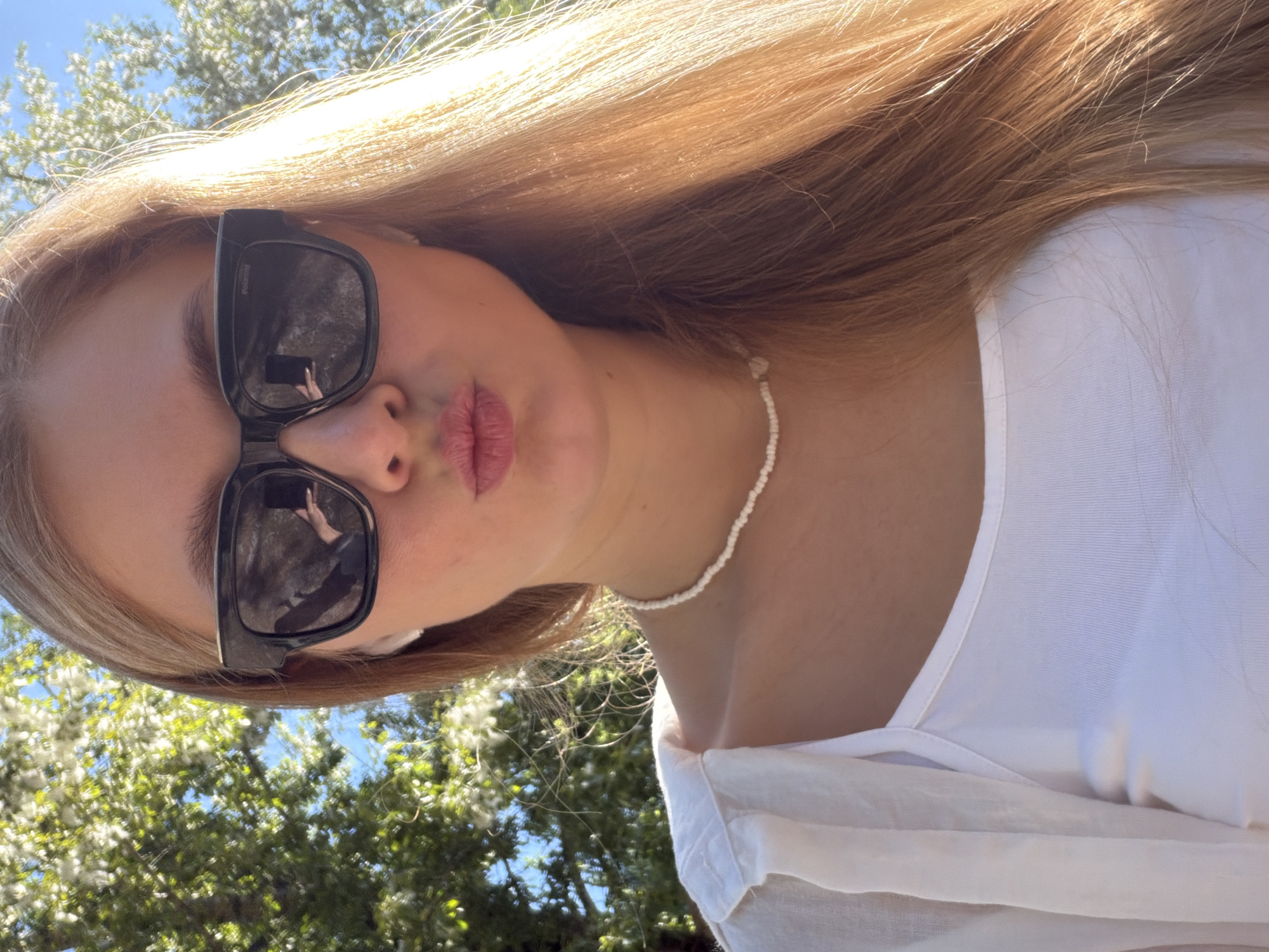 A young woman with long blonde hair and a white top, wearing black sunglasses and a white beaded necklace, poses for a selfie outdoors. She is puckering her lips and has white earbuds in. In the background, green trees with white fluffy seed pods are visible against a blue sky. The lighting suggests it is daytime. There is no information to identify the location as Chaikovsky, Russia.