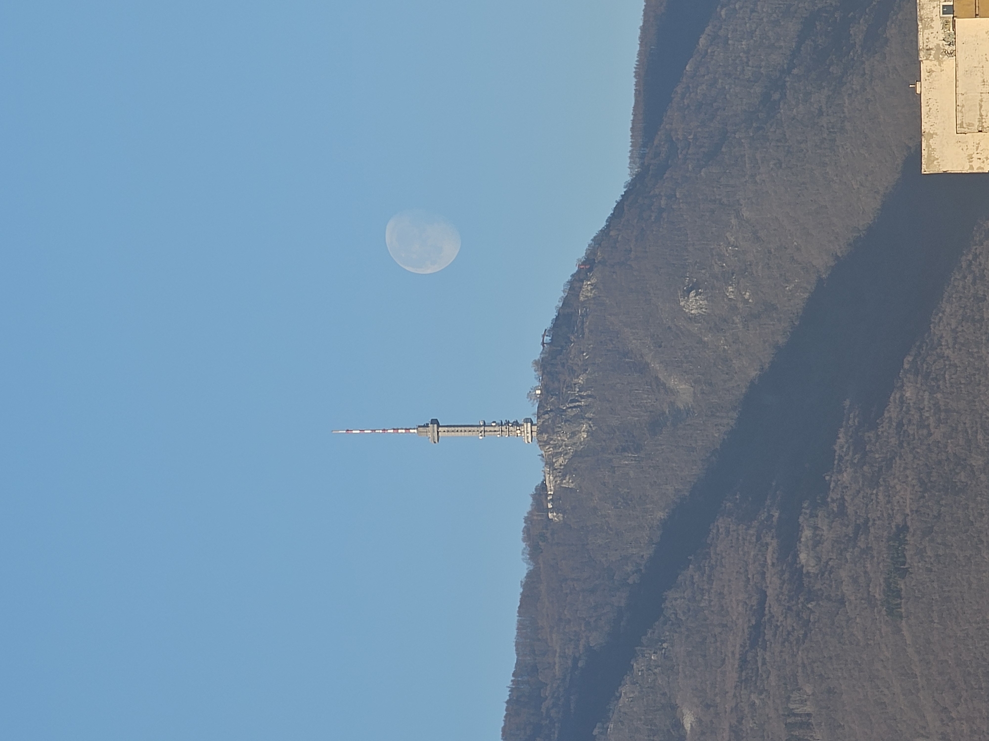 The image depicts the Kopitoto TV Tower on Vitosha Mountain, near Sofia, Bulgaria, with a half moon visible in the blue sky above the tower. A forested mountain slope occupies the right side of the frame, while a building is visible at the top right corner.