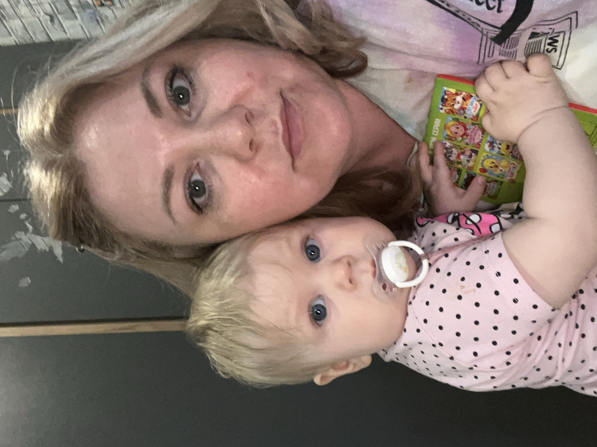 A woman and a baby are positioned close to each other. The woman has blonde hair and fair skin, with evident makeup around her eyes. The baby, also fair-skinned with blonde hair, has a pacifier in its mouth and is wearing a pink polka dot top. The baby is holding a colorful children's book. The background appears to be a dark, solid color, possibly a wall. The photograph is taken from a close, slightly overhead angle. There is no evidence or information to determine the location in the city of Vesyoloye, Russia.