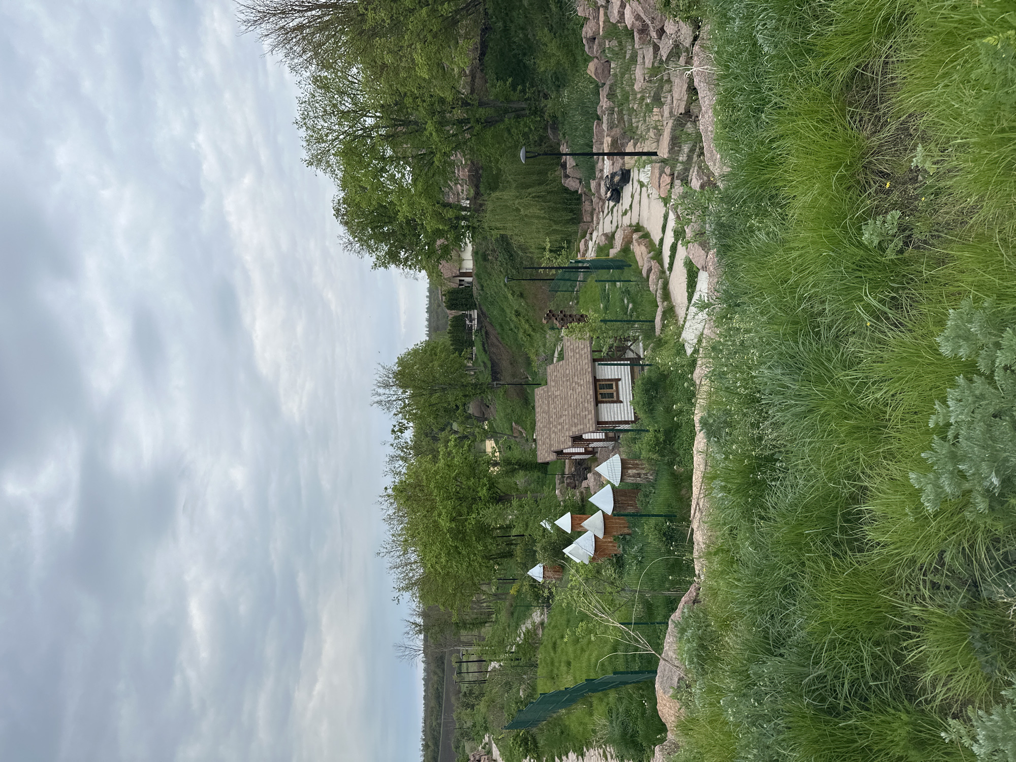 The image shows a landscape in Zakharivka, Ukraine, featuring a winding stone path lined with grass and rocks. A small house with a brown roof is visible amidst trees. There are several white triangular objects, possibly decorations, on a green fence. The sky is overcast with gray clouds. No people are visible in the scene.