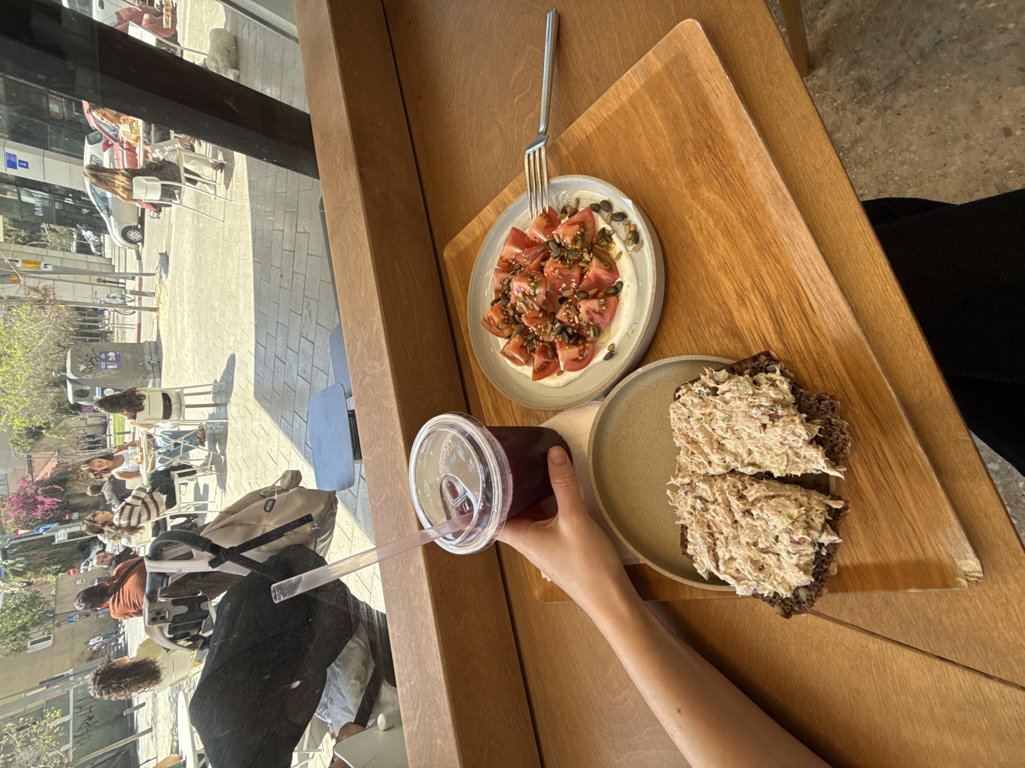 The photograph captures a moment inside a cafe in Tel Aviv, Israel. The scene is viewed from an elevated perspective looking out a window. Outside the window, a sunlit sidewalk bustles with activity. People are seen sitting at tables, walking, and pushing strollers.
Inside the cafe, a wooden tabletop is the central focus. On the table, a rectangular wooden tray holds two plates and a beverage. One plate contains a salad of chopped tomatoes with seeds. A fork rests next to this plate. The second plate holds two slices of dark bread topped with a salad of light-colored salad. A hand is holding a plastic cup containing a dark-colored beverage, with a straw visible. The photograph is taken on a sunny day, casting light both inside and outside the cafe.