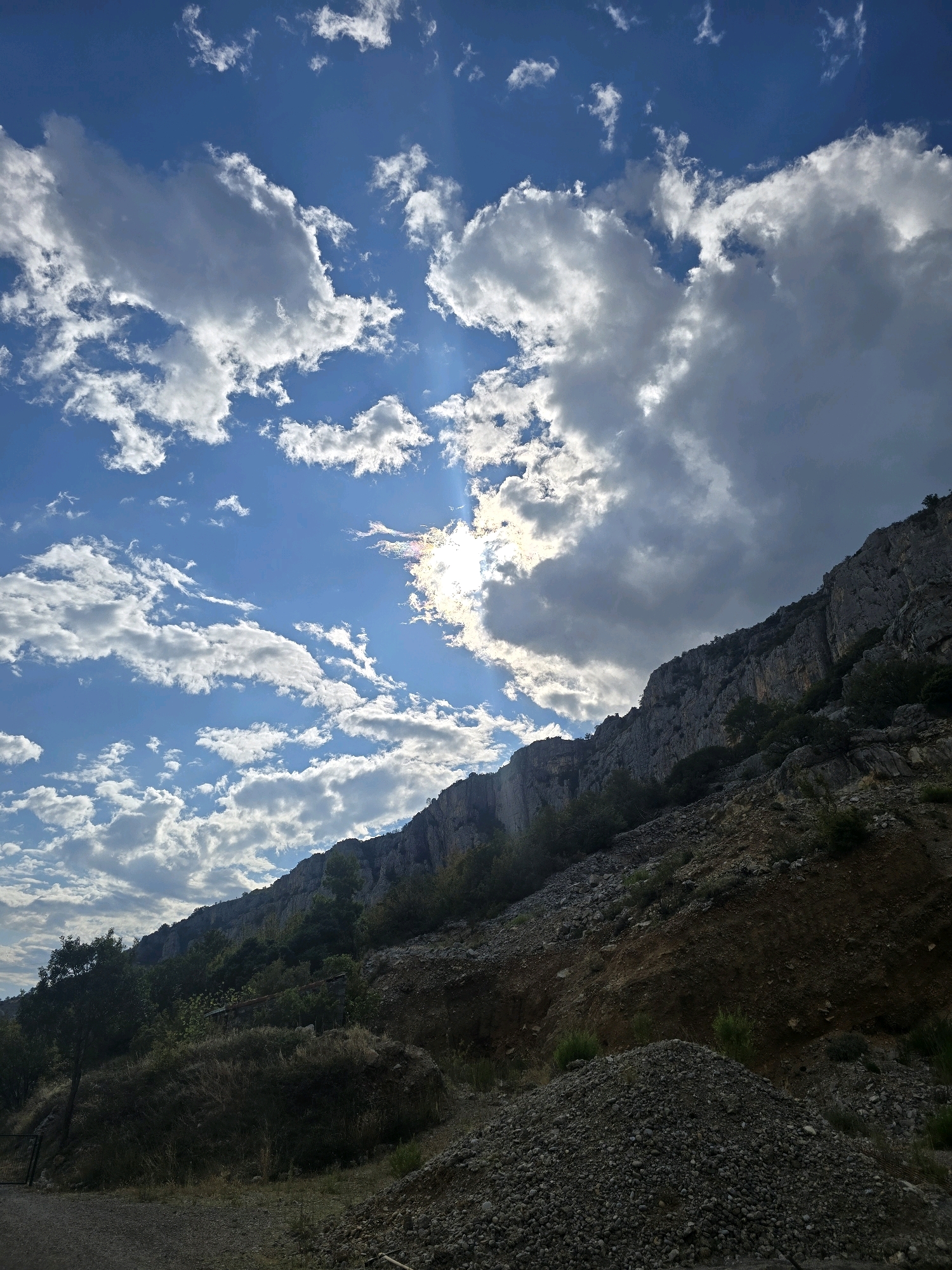 The image depicts a view of a rocky hillside with sparse vegetation against a bright blue sky filled with dramatic white and gray clouds. Sunlight pierces through the clouds in some areas, creating a strong contrast. The hillside is rugged, with visible layers of rock and scree. In the foreground, there are piles of gravel and dry grass. The terrain suggests an undeveloped or natural area, possibly on the outskirts of a city. The scene is likely located in Podgorica, Montenegro, given the characteristic karst landscape and vegetation found in the region. There are no people or animals visible in the image.
