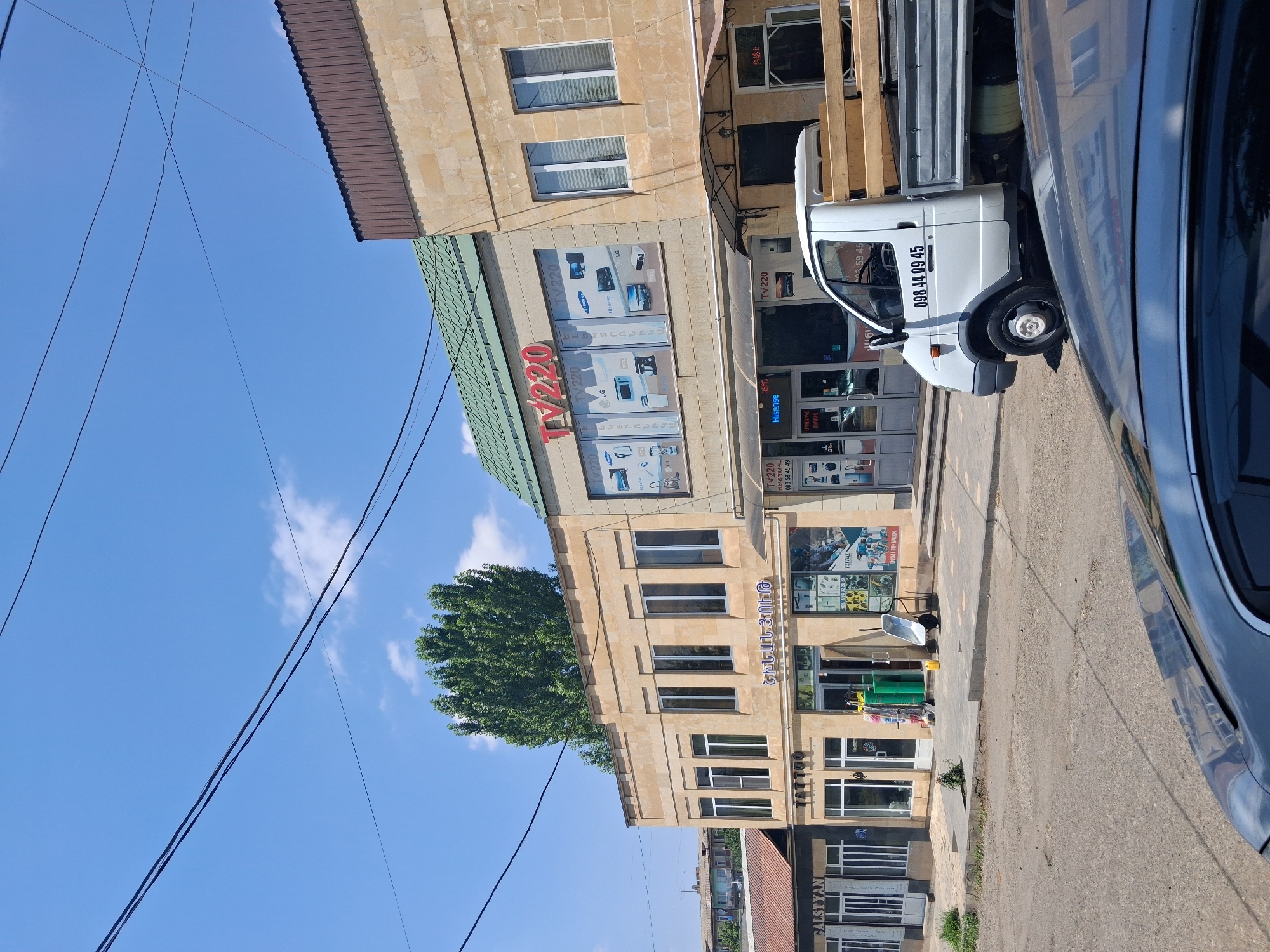 The image depicts a street scene in Yeghegnadzor, Armenia, on a clear, sunny day. In the foreground, the hood of a dark-colored car reflects the surrounding environment. To the right, a white flatbed truck is parked, its bed loaded with wooden planks. The truck has a license plate with the number "098 44 09 45" prominently displayed on its side.
Behind the truck are several commercial establishments. The most prominent is a building with a green tiled roof and a beige stone facade. This building houses a store with a large sign reading "TV220." The store's windows display various electronic products, including televisions and computers, with brand names like LG, TCL, Samsung, and Hisense visible. Adjacent to this is another store with a beige facade and large windows, featuring signage for "TATTOO." Further to the left, a building with a brown tiled roof and a beige stone facade has a sign that reads "GALSTYANI."
The street in front of the stores is paved with asphalt and concrete. There is a sidewalk area with some green patches of grass. In front of the "TV220" store, there are several green cylindrical objects, a yellow bucket, and a wheelbarrow. Overhead, power lines crisscross the bright blue sky, which is dotted with a few white clouds. A large, leafy green tree is visible behind the buildings, and in the distance, other buildings with red tiled roofs can be seen. The overall impression is that of a commercial street in a town or city.