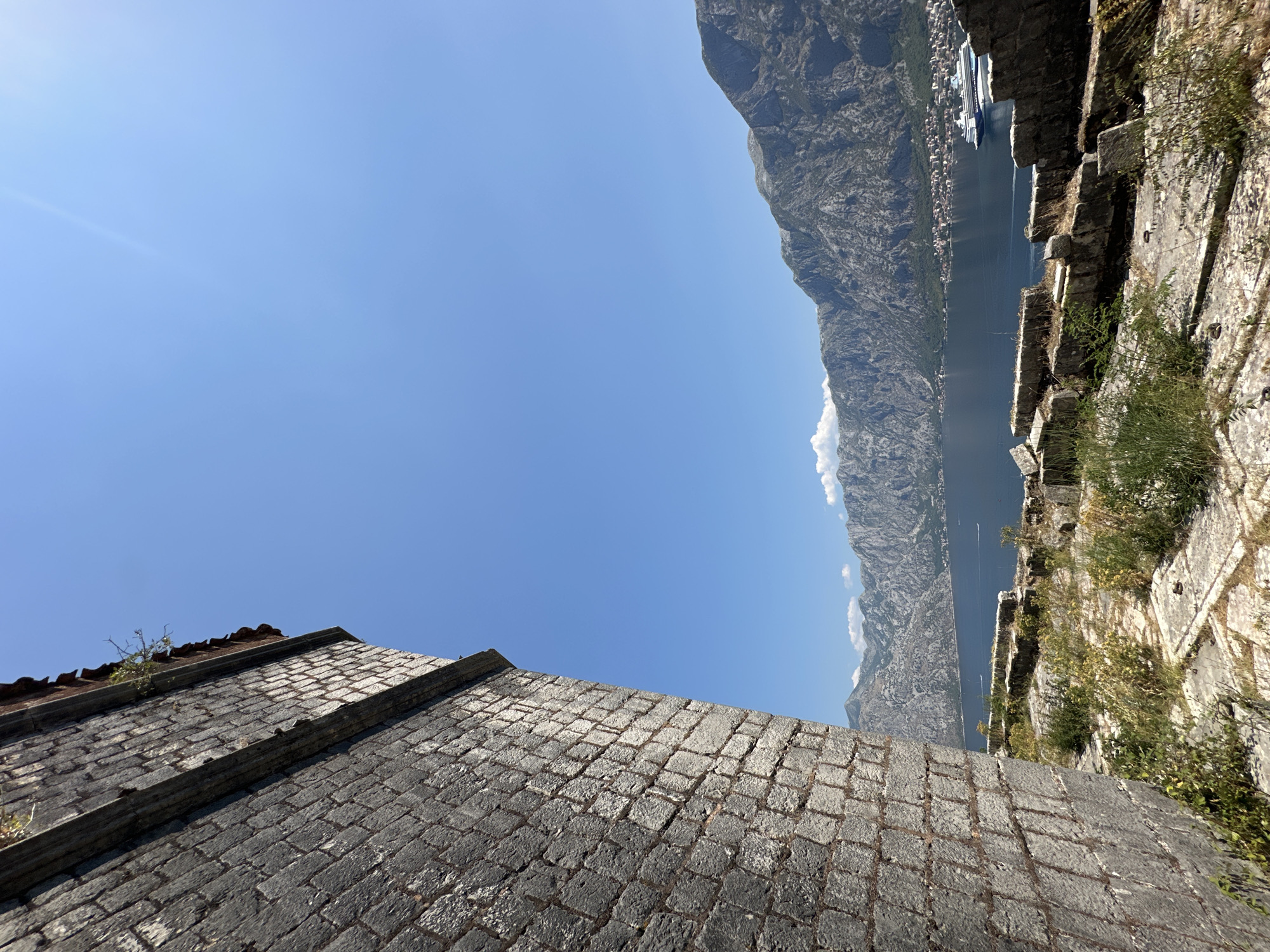 A view from a high vantage point within a stone structure in Dobrota, Montenegro, overlooks a bay and distant mountains. In the foreground, a weathered stone wall, constructed of rectangular blocks with visible mortar lines, occupies the left side of the frame. The wall angles into the image from the left. Beyond the wall, the ground is covered in stone slabs and patches of dry grass and small plants, suggesting a somewhat overgrown and ancient area.
The middle ground features a wide expanse of dark blue water, likely a bay or fjord, with several small white wakes from unseen vessels. A large, white cruise ship with blue accents is visible on the water, moving from left to right.
The background is dominated by a series of majestic, rugged mountains that rise sharply from the coastline. A small town or settlement with red-tiled roofs is nestled along the edge of the water at the base of the mountains. The sky is a clear, bright blue with a faint lens flare visible in the upper right corner. The overall impression is one of historical grandeur and natural beauty under a sunny sky.