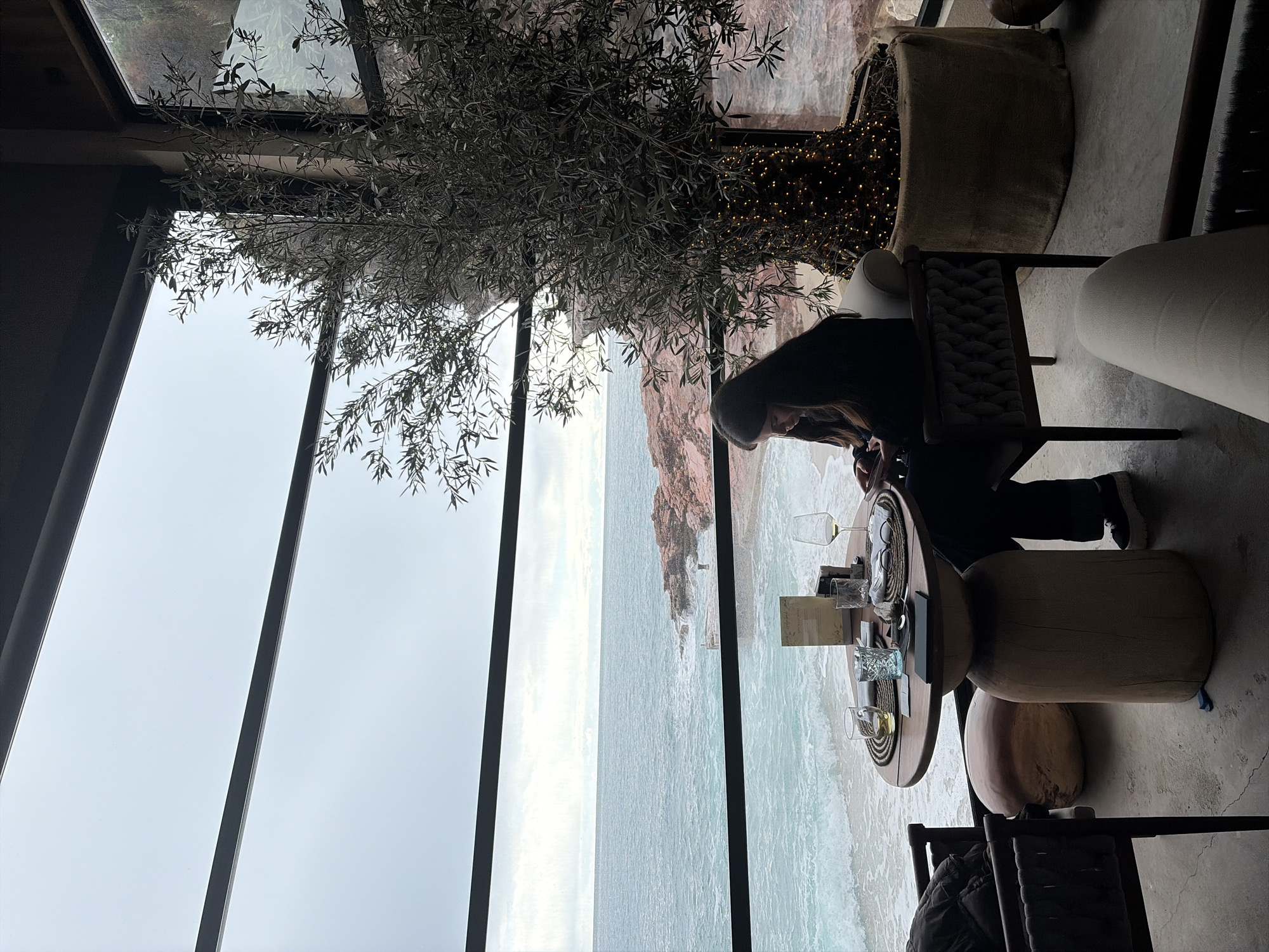 The scene depicts the interior of a coastal restaurant or cafe located in Unknown, Unknown, with a large window offering an unobstructed view of the ocean. A female individual with long dark hair, wearing a dark top and dark pants, is seated at a round wooden table, focused on a small electronic device in her hands. The table is set with two woven placemats, a menu, a wine glass containing white wine, a glass with water, another glass with an amber liquid, and cutlery. To the right of the table, a large potted tree, resembling an olive tree, with its trunk wrapped in warm-toned string lights, stands beside the window. The window frames a view of a light blue-green ocean with breaking waves crashing against a reddish-brown rocky shoreline under an overcast, grey sky. A small white structure and a solitary figure are visible on the distant rocks. The interior features light grey concrete flooring, and various modern chairs and a large, rounded, light-brown cushion are positioned within the space.