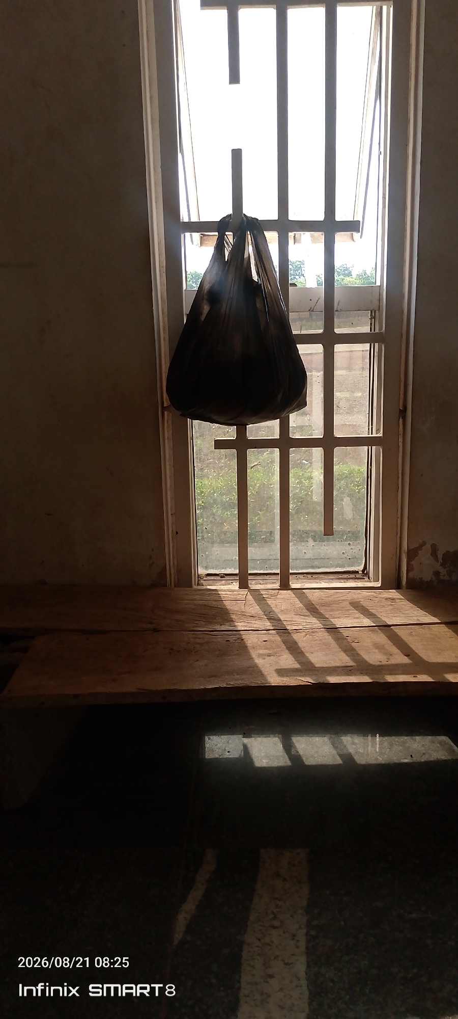 A plastic bag hangs from a white metal bar in front of a window in Kakau, Nigeria. The bag is dark and appears to contain items. The window is covered with white bars and the glass is dirty. Sunlight streams through the window, casting shadows of the bars on a wooden surface below. The wooden surface is a plank of wood, possibly a makeshift table or shelf, positioned in front of the window. The floor below is dark and reflects the light and shadows from the window. The walls are plain and appear to be made of concrete. A timestamp at the bottom left corner reads "2026/08/21 08:25" and "Infinix SMART 8". The scene suggests an interior setting with a view to the outside through a barred window.