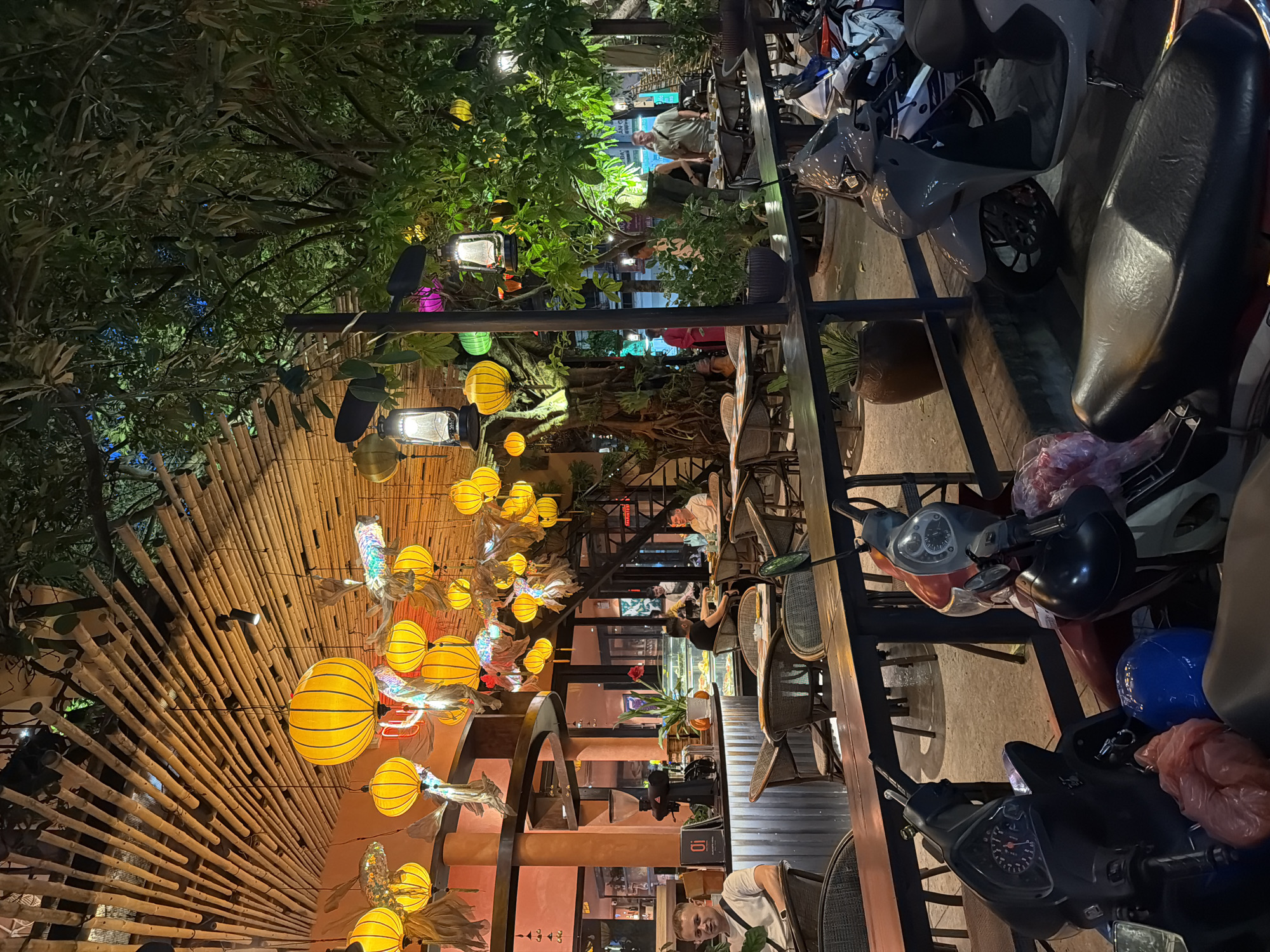 The image depicts an outdoor seating area of a cafe or restaurant at night, located in Nha Trang Ward, Vietnam. The space is illuminated by numerous hanging yellow lanterns and a few electric lamps, creating a warm and inviting atmosphere. The ceiling is constructed from bamboo poles arranged in a radial pattern.
In the foreground, several motorbikes are parked. The seating area features a long, dark wooden table with chairs and tables set up in front of a building. The building's facade appears to be painted in an orange hue, with large windows revealing the interior. Inside, a counter with a coffee grinder and display cases with food are visible. Several people are seated at tables, engaged in conversation or dining.
To the right of the seating area, a large tree with dense foliage is prominent, with more lanterns hanging from its branches. The overall impression is a bustling yet relaxed evening scene in a popular urban establishment. The ground is paved, and there are potted plants contributing to the ambiance. The presence of motorbikes suggests a common mode of transportation in the area.