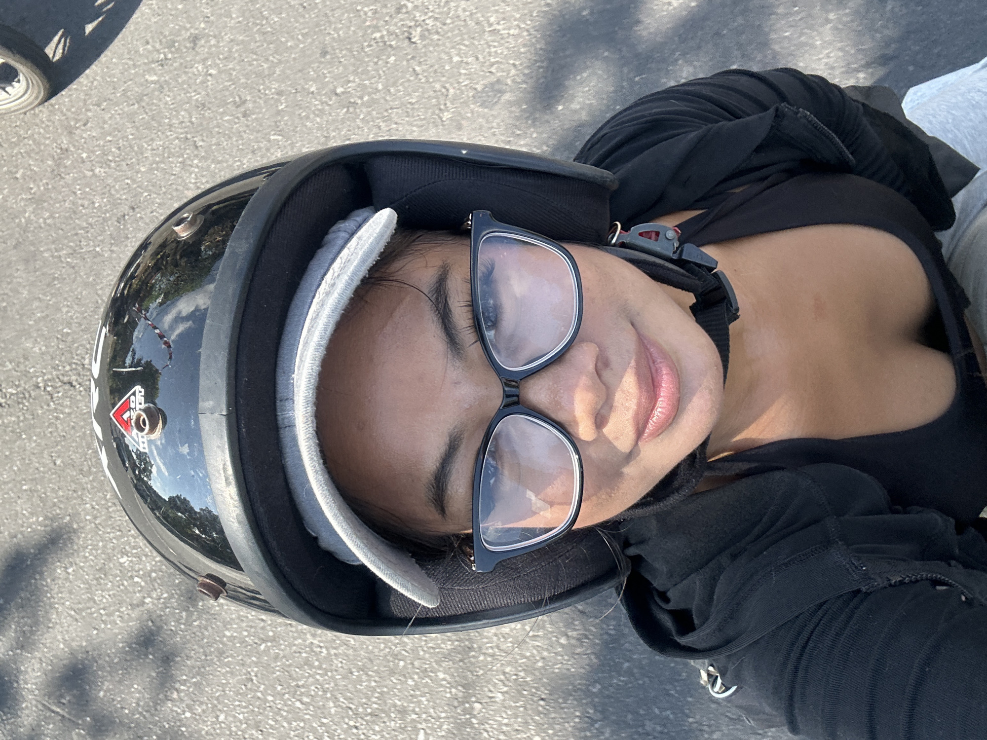 A woman is riding a motorcycle in Sukawati, Indonesia. She is wearing a black helmet, a gray baseball cap, and glasses. She is also wearing a black hoodie over a black top. The road is made of asphalt and there are shadows of trees on the ground. The woman is looking at the camera and smiling.