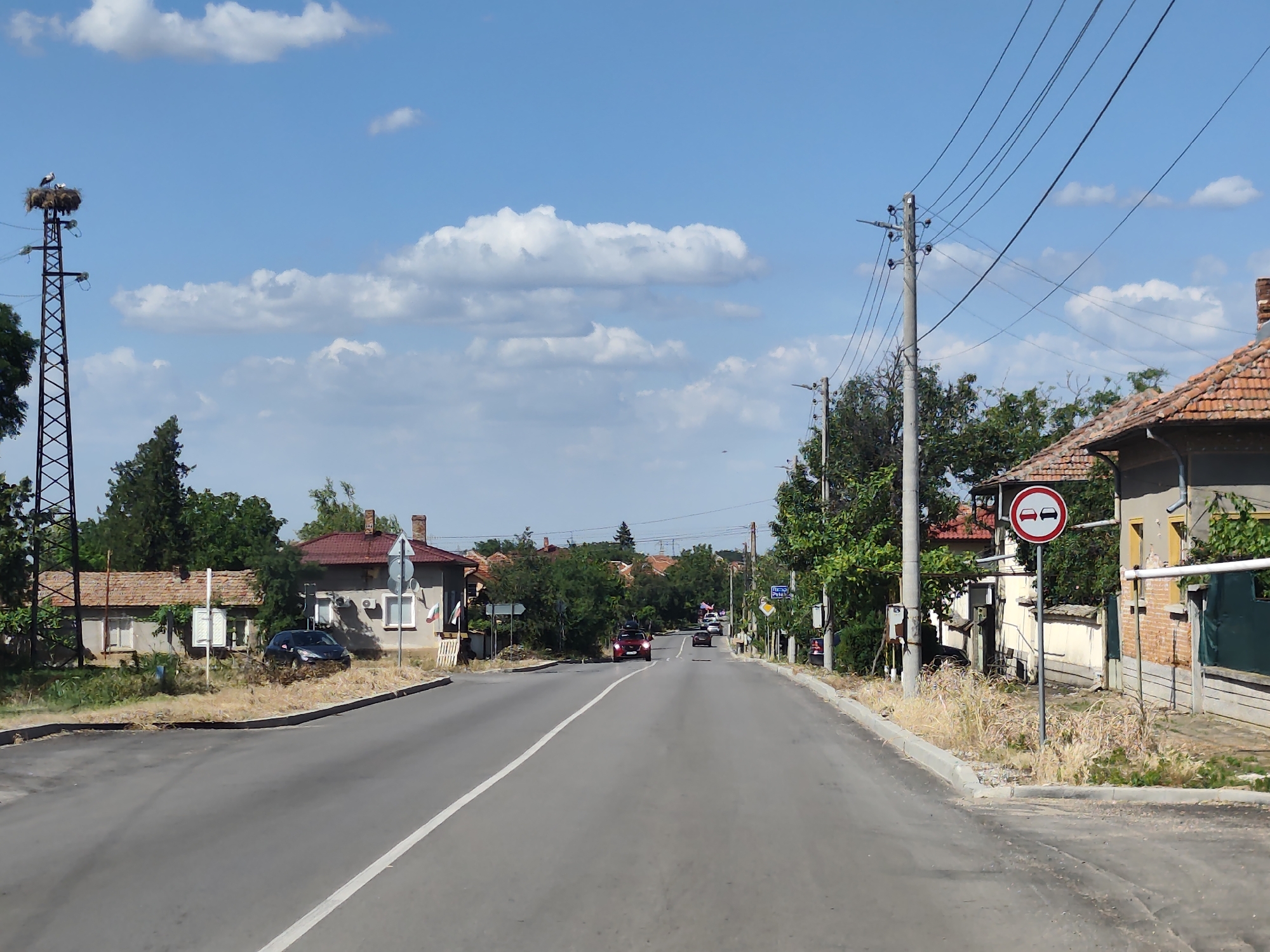 This is a forward-facing, eye-level, medium shot photograph taken on a sunny day in Krushovitsa, Bulgaria. The scene depicts a two-lane road lined with houses and trees, leading into a village. The sky is a clear blue with scattered white clouds.
On the left side of the road, a tall, metal electrical tower supports a stork's nest with two storks. Further down the road, a dark blue car is parked in front of a small house. On the right side of the road, a traffic sign shows two cars with a red circle and a red line through them, indicating "no overtaking." Beyond this, more houses and trees are visible.
Several cars are driving on the road, heading away from the camera. The overall mood is peaceful and rural. The time of day appears to be late morning or early afternoon, given the bright sunlight and clear sky. The vegetation suggests a warm season, likely summer. There is some dry grass along the side of the road, indicating dry conditions. A street sign with Cyrillic text is visible on the right side of the road in the distance.