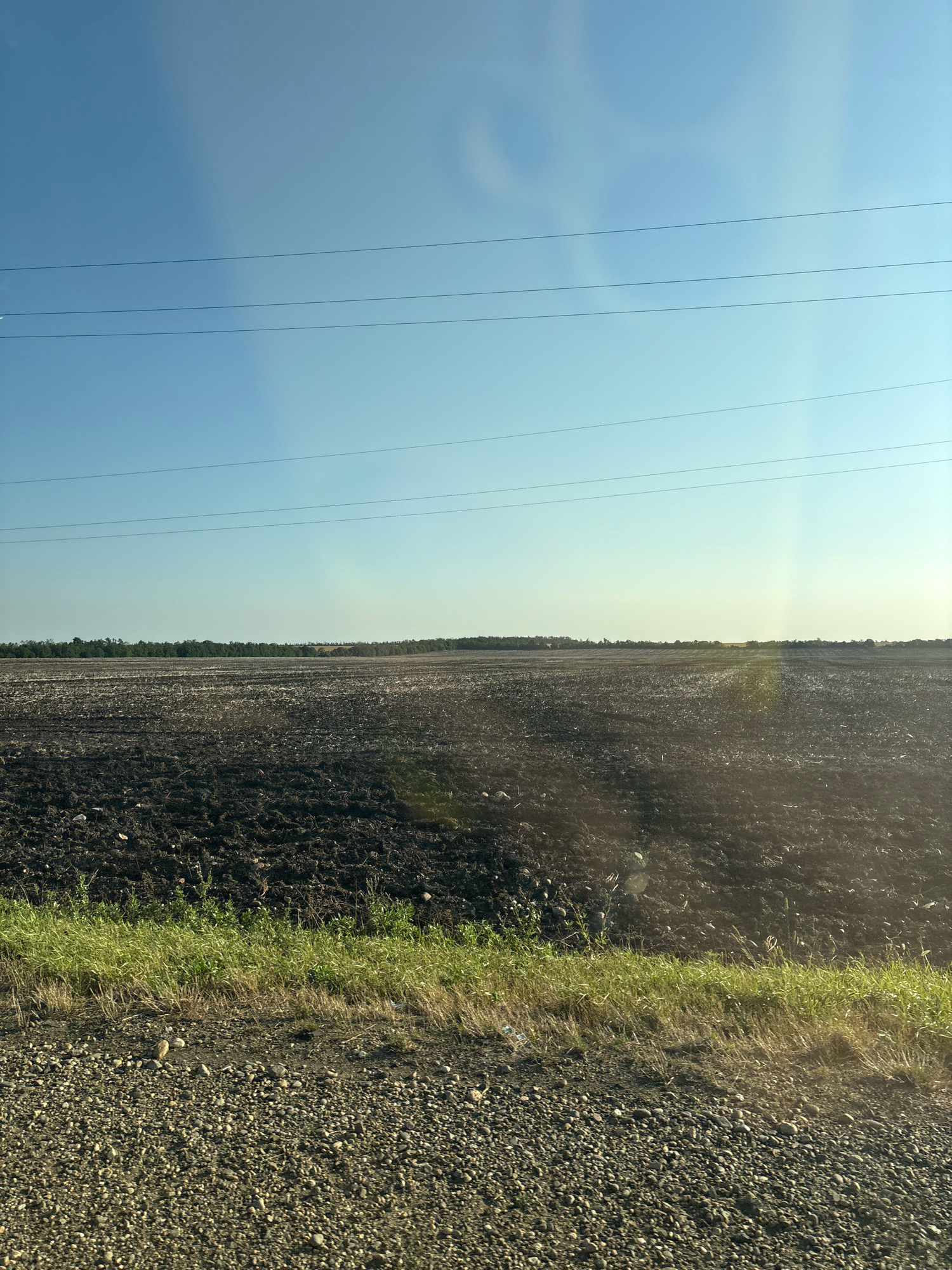 A wide, plowed field under a clear blue sky dominates the image. Power lines stretch horizontally across the sky in the upper portion of the frame. The field, appearing dark and freshly tilled, stretches to the horizon where it meets a line of trees. The foreground consists of a gravel and dirt embankment with some sparse green grass. The scene appears to be located in a rural area, likely Glivenko, Russia, given the context. There are no people or animals visible in the image. The overall impression is one of agricultural land, possibly prepared for planting or recently harvested.