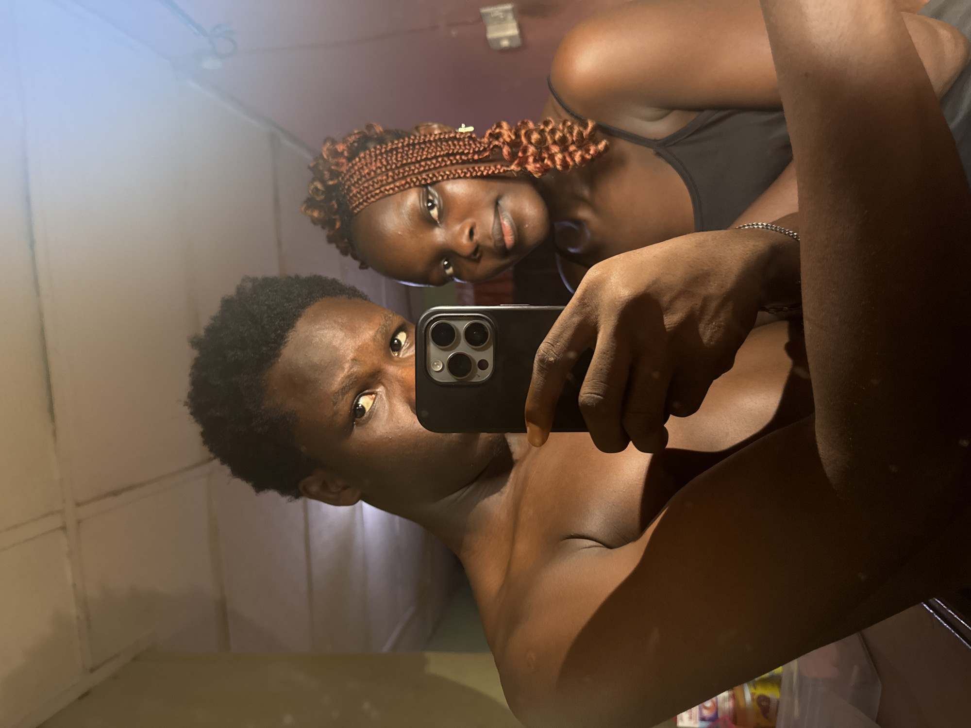 In a dimly lit room, a young man and woman pose for a mirror selfie. The man, shirtless, holds a smartphone in his right hand, capturing their reflection. His left eye is visible, looking directly at the camera, while the rest of his face is partially obscured by the phone. The woman stands beside him, also looking towards the camera. She has braided hair with reddish-brown extensions and wears a black tank top. The background reveals a textured ceiling and a plain wall. The overall ambiance suggests an informal setting within a residential space in Onitsha, Nigeria, possibly a bedroom or living area.