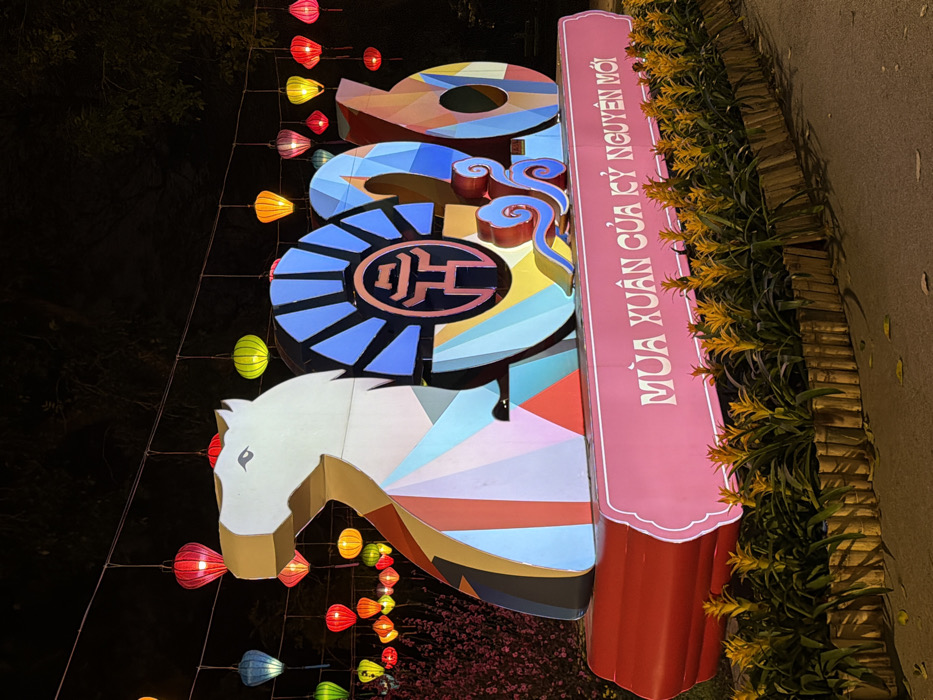 The image displays a vibrant, illuminated public art installation at night in Hà Nội, Vietnam, likely situated in a prominent festive area such as around Hoàn Kiếm Lake or a central city park. The central feature is a large, freestanding structure spelling out '2026'. The first numeral '2' is stylized as a horse head, primarily white, with geometric multi-colored segments (blue, pink, brown, red) forming its lower body. The '0' is a blue, spoked wheel-like form, containing a central dark emblem outlined in red, resembling a traditional Vietnamese character or crest. The subsequent numerals '2' and '6' are composed of angular, multi-colored panels. This entire structure rests on a red base, which is fronted by a pink plaque bearing the white Vietnamese text 'MÙA XUÂN CỦA KỶ NGUYÊN MỚI' (Spring of the New Era). In the foreground, a landscaped bed contains rows of yellow-orange flowering plants, enclosed by a low border constructed from vertical wooden segments. Overhead, numerous traditional Vietnamese lanterns in assorted colors—red, yellow, green, blue, orange, and pink—are suspended from wires, providing ambient illumination. Blurred pink blossoms are discernible in the left background.