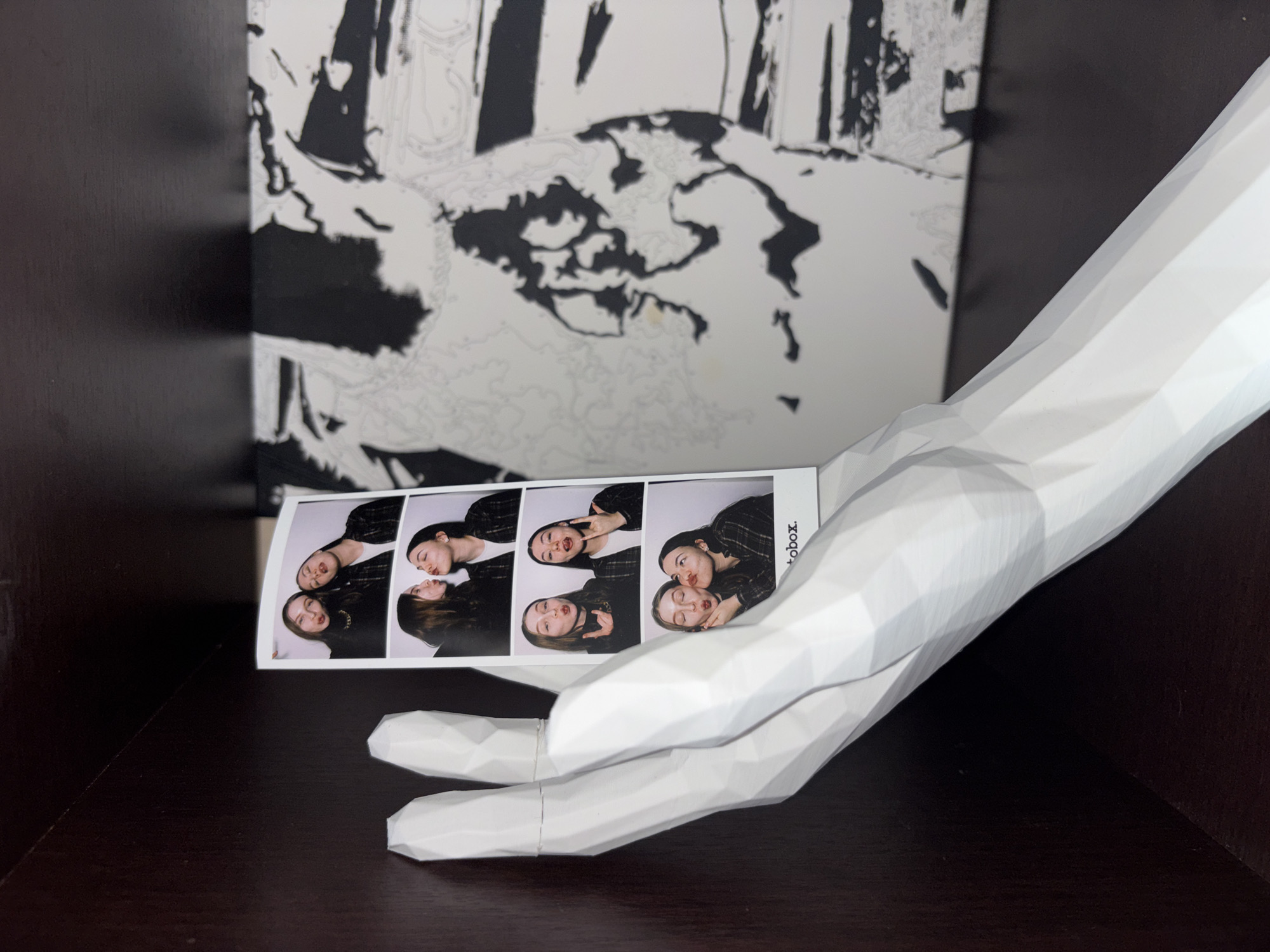 The image depicts a dark brown shelf or surface on which a white, polygonal hand-shaped sculpture holds a photobooth strip of two young women in casual clothes. The photobooth strip contains four images of the women posing together in various playful ways. Behind the hand sculpture and photobooth strip is a black and white abstract art piece. The location in Perm, Russia is unknown.