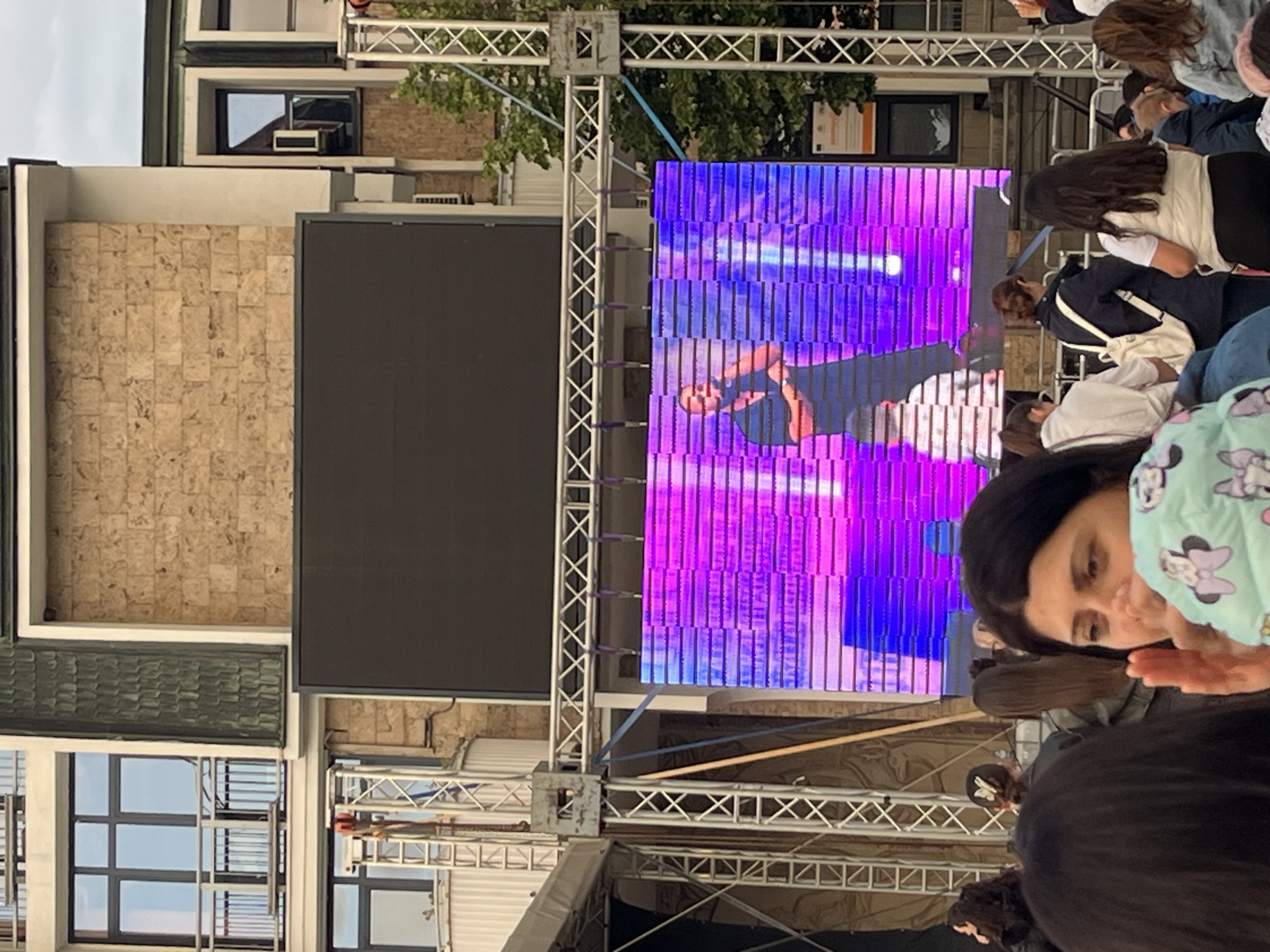 Here is a detailed description of the image:
**Scene:** The image depicts a public event held outdoors, possibly a concert or festival, taking place in an urban setting. The event features a large screen displaying a performer on stage. The crowd is visible in the foreground. Buildings are present in the background.
**People:**
* A crowd of people is gathered to watch the performance on the screen.
* A woman with long dark hair is partially visible in the foreground. She wears a jacket. A child in a light green jacket with Mickey Mouse design on it is also in front.
* The performer is shown on the screen.
**Objects:**
* Large LED screen mounted on a scaffolding structure.
* The screen is displaying a performer under stage lighting.
* Buildings are visible in the background. One of them features a tiled facade.
* Windows are visible on the buildings.
* Trees/foliage visible above and to the sides of the screen.
**Environment:**
* Outdoor urban environment.
* Daytime, with cloudy skies.
* Buildings in the background indicate a city setting.