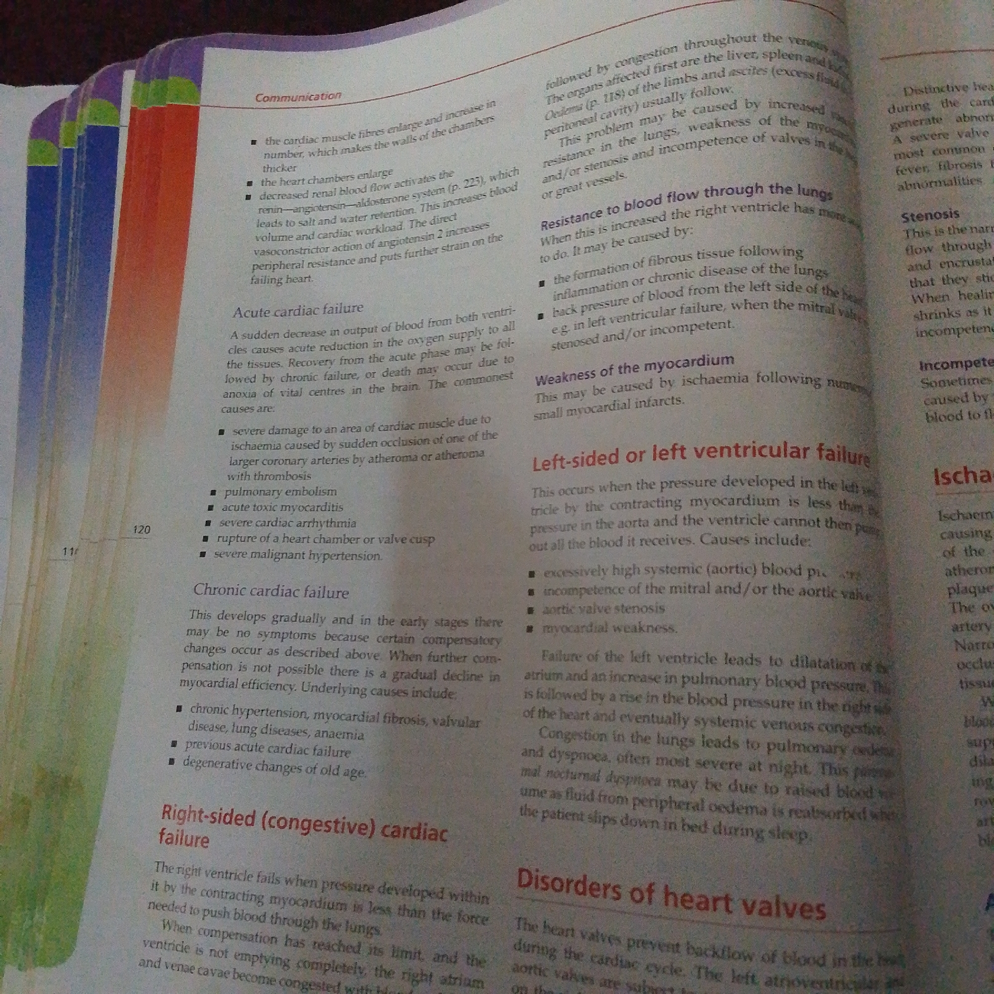The image is a close-up, overhead shot of an open book, showing two pages of text with colorful page edges. The book appears to be a medical or biology textbook, focusing on the heart and its failures. The text discusses various types of cardiac issues, including "Acute cardiac failure," "Chronic cardiac failure," "Left-sided or left ventricular failure," and "Disorders of heart valves." Specific medical terms like "myocardium," "ischaemia," "atheroma," and "stenosis" are visible. There are bullet points and numbered lists within the text, indicating structured information. The page edges are brightly colored, with stripes of blue, green, red, and orange, suggesting they might be dividers or visual cues within the book. The overall setting is a flat surface, likely a table or desk, where the book is laid open. There are no people or distinct environmental features visible, only the book itself and the text it contains. The lighting appears to be artificial, illuminating the pages clearly.