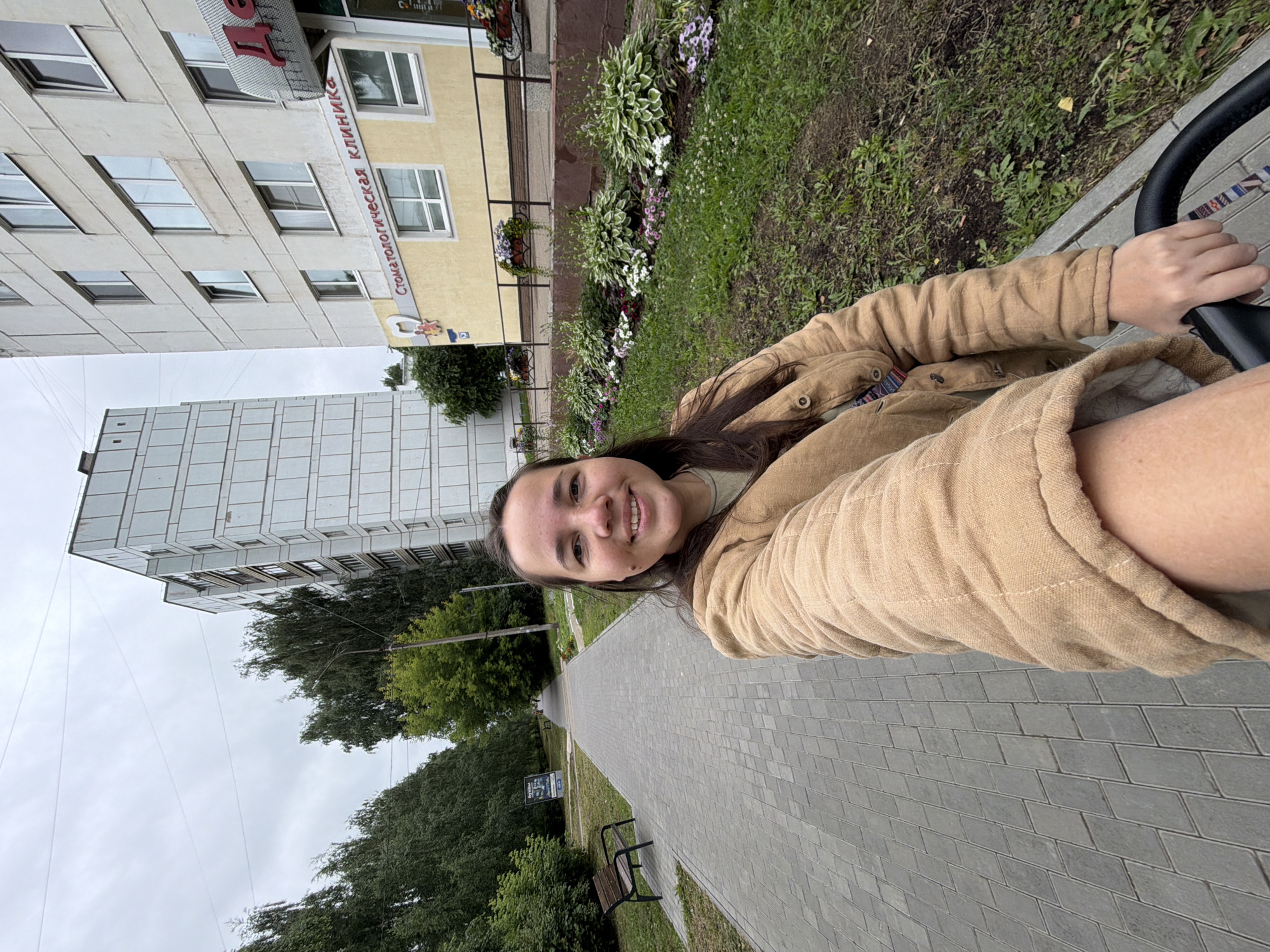A woman with dark hair and a smile takes a selfie outdoors in Chaikovsky, Russia. She is wearing a tan jacket and is holding a stroller. The stroller's handle is visible in the lower right corner. The background shows a paved walkway, a row of trees, a tall building, and a building with a sign that reads "Стоматологическая клиника" (Dental Clinic). There are flower beds in front of the dental clinic. The sky is overcast.