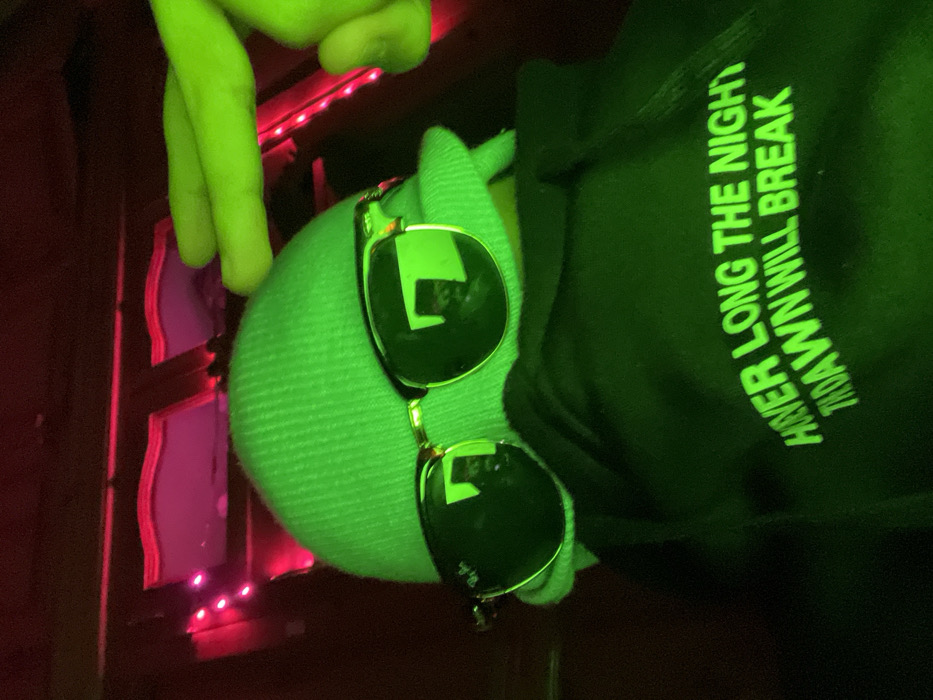 A lime green plush Kermit the Frog doll wearing sunglasses sits on a dark surface. The doll is in the lower middle of the frame, with its legs dangling above. The background consists of a dark structure featuring red and pink lighting elements. The doll is also in front of a black hoodie with glowing text saying, "HOVER LONG THE NIGHT TILL DAWN WILL BREAK". The image is taken at an unusual angle.