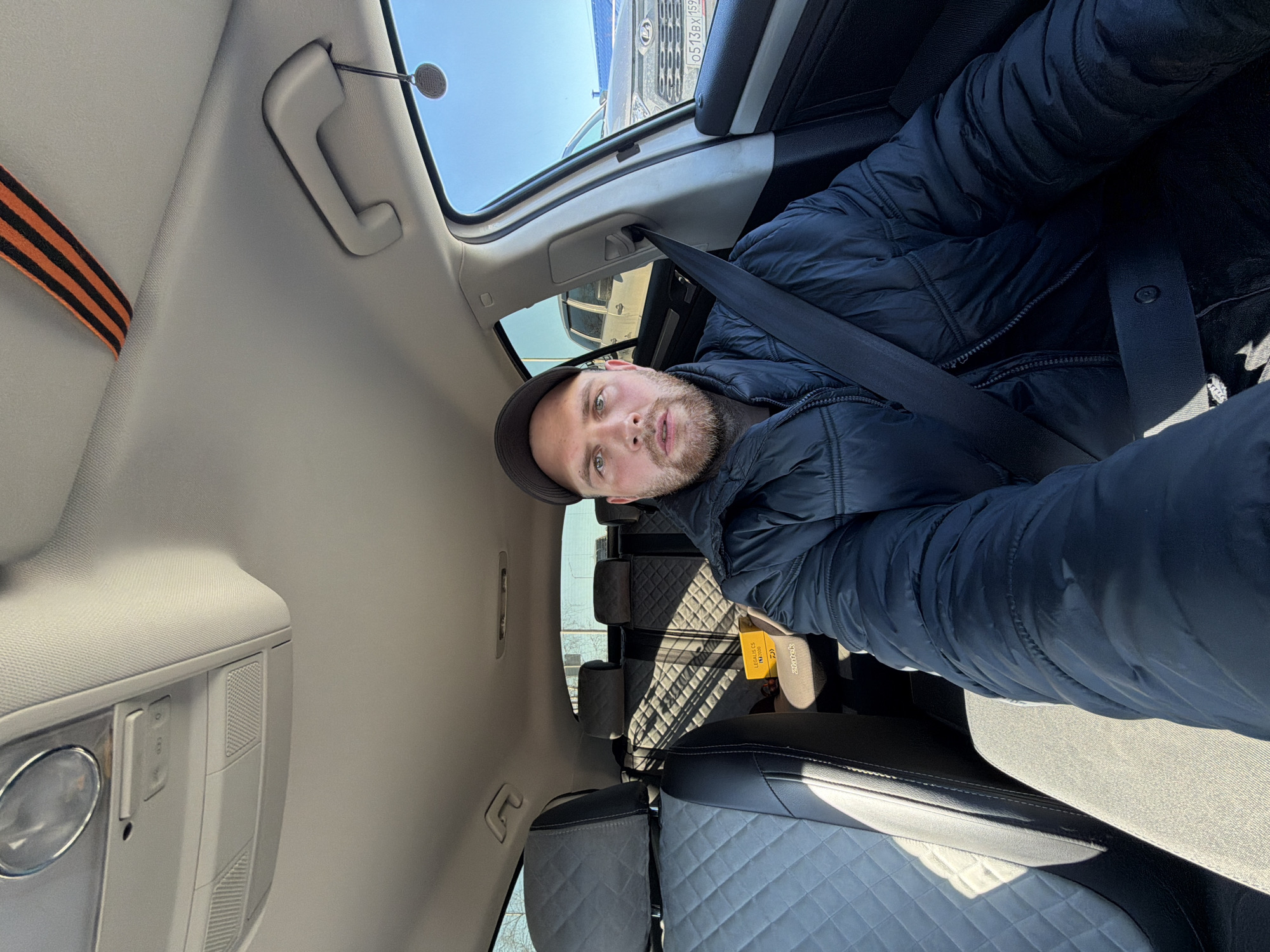A man is seated in a car, wearing a black puffer jacket and a dark cap. He has a beard and fair skin. The safety belt is fastened. The interior of the car is light gray with gray seats with a quilted pattern. In the back seat, there's a package of fishing line on the floor. A black and orange striped ribbon is visible above the man's head, attached to the interior of the car near the roof. Outside the car, a light-colored truck is parked. Based on the license plate O 513 BX 159 visible on the truck, the location is Chaikovsky, Russia. The sky is clear and blue. The image is a selfie taken from inside the car.