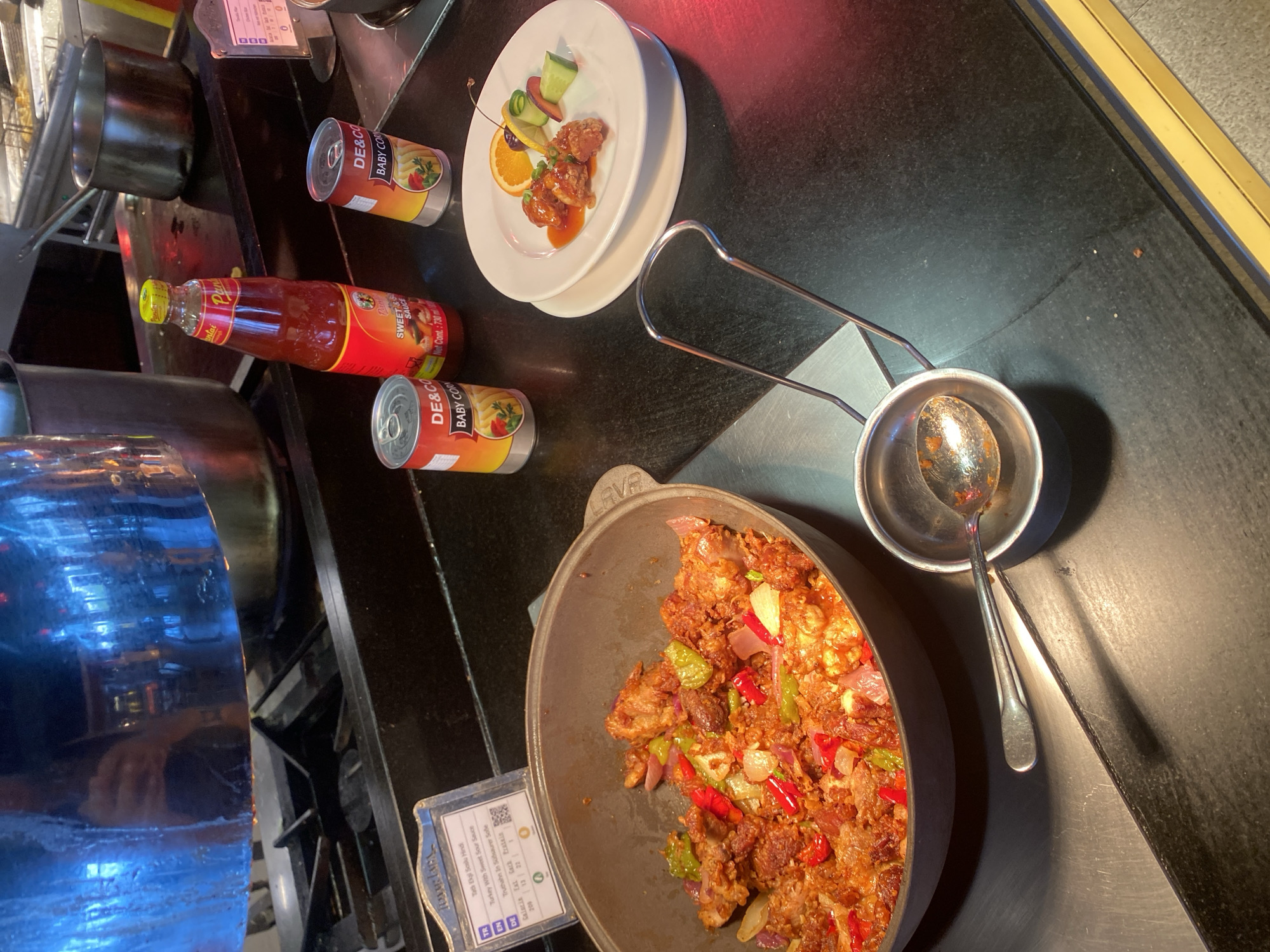The image shows a buffet-style food presentation. In the foreground, a large dark gray pot contains a dish with what appears to be fried chicken pieces mixed with red and green peppers, and red onions. A metal serving spoon rests in a small metal bowl next to the pot. To the right, a white plate holds three pieces of glazed food, garnished with an orange slice, a cherry, and cucumber.
In the background, two cans of "DE&CO BABY CORN" are visible, along with a bottle of red sauce labeled "Pantai Sweet & Sour Sauce". A metal pot is positioned behind these items. A reflective metal surface with blue lighting is above the stove. A small informational sign is placed to the left of the pot, with text including "Turkey" and "Sweet Sour Sauce".
The setting appears to be a restaurant or buffet area. There is no visible indication of the city of Aksu, Turkey, in the image.