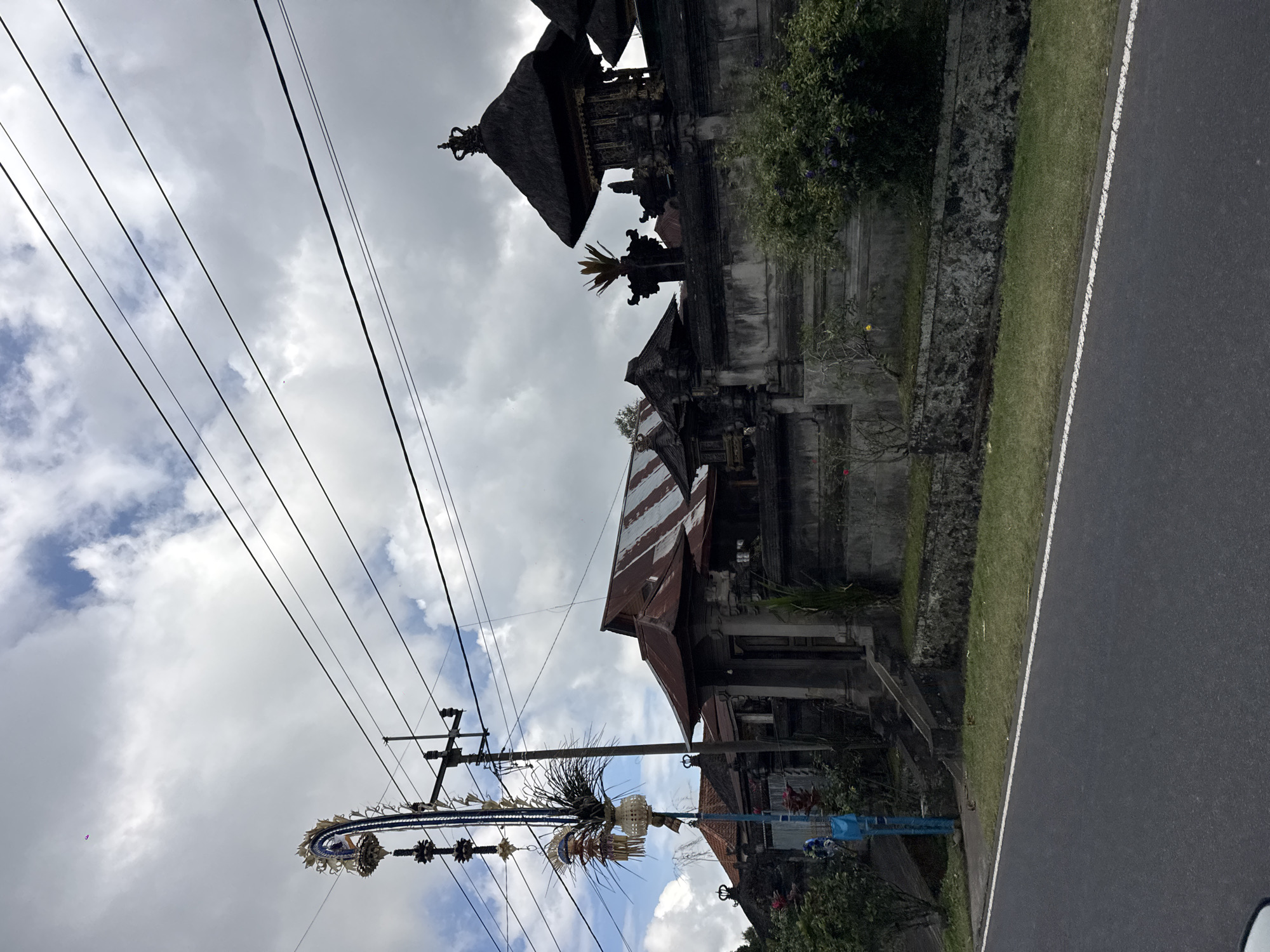 The image depicts a street scene in Susut, Indonesia, with a roadside temple or traditional Balinese building on the left. The building features multiple tiers with thatched roofs and ornate carvings. Power lines crisscross the sky, and a tall pole adorned with elaborate, woven decorations hangs prominently in the foreground. The decorations appear to be festive and traditional, possibly related to a local celebration or offering.
Adjacent to the temple is a grassy verge and a paved road with a white line marking the edge. Lush green trees and foliage are visible around the temple complex. The sky is filled with dramatic, cloudy formations, suggesting either an overcast day or the aftermath of a storm. The overall atmosphere is somewhat subdued due to the weather, but the intricate decorations hint at vibrant cultural practices. No people are clearly visible in the frame. The time of day is not explicitly indicated, but the lighting suggests it could be morning or late afternoon. The image is taken from a low angle, looking upwards towards the sky and the structures.