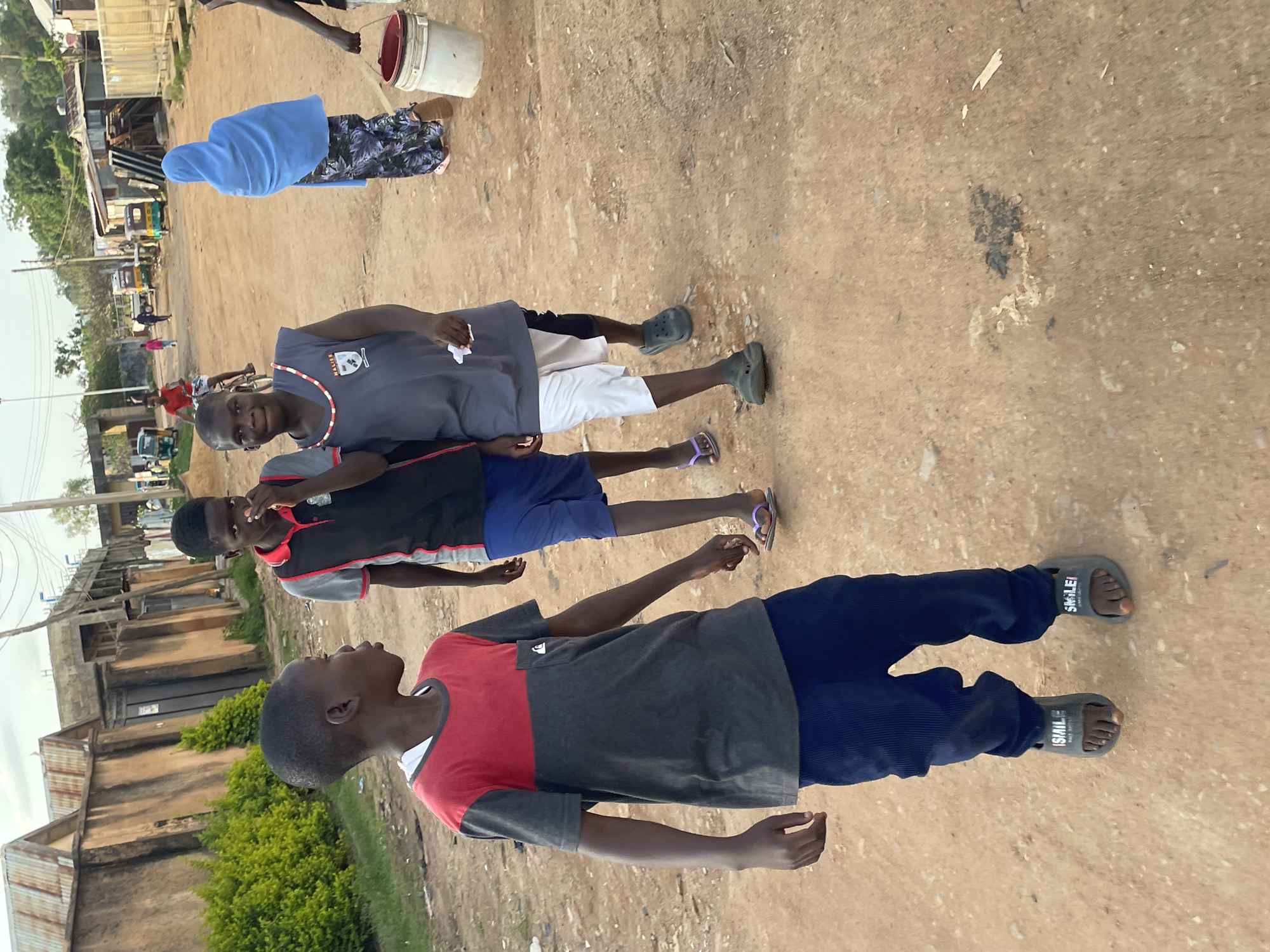 In Barakin Akawo, Nigeria, four young people are captured walking on a dirt road under a slightly overcast sky. In the foreground, a boy wearing a red and grey t-shirt and dark blue pants walks towards the camera, his feet clad in grey slides with "SMILE" written on them. To his right, another boy, dressed in a black and red t-shirt and blue shorts, is seen with his hand near his mouth. Further back and to the right, a third boy in a grey sleeveless shirt with white and black shorts walks, holding a white paper. Behind him, a woman in a blue hijab and a patterned skirt carries a white bucket. The background reveals buildings with corrugated metal roofs, some appearing unfinished. A few vehicles, including a yellow tuk-tuk and a white truck, are visible in the distance. The ground is a mix of loose dirt and some scattered debris.