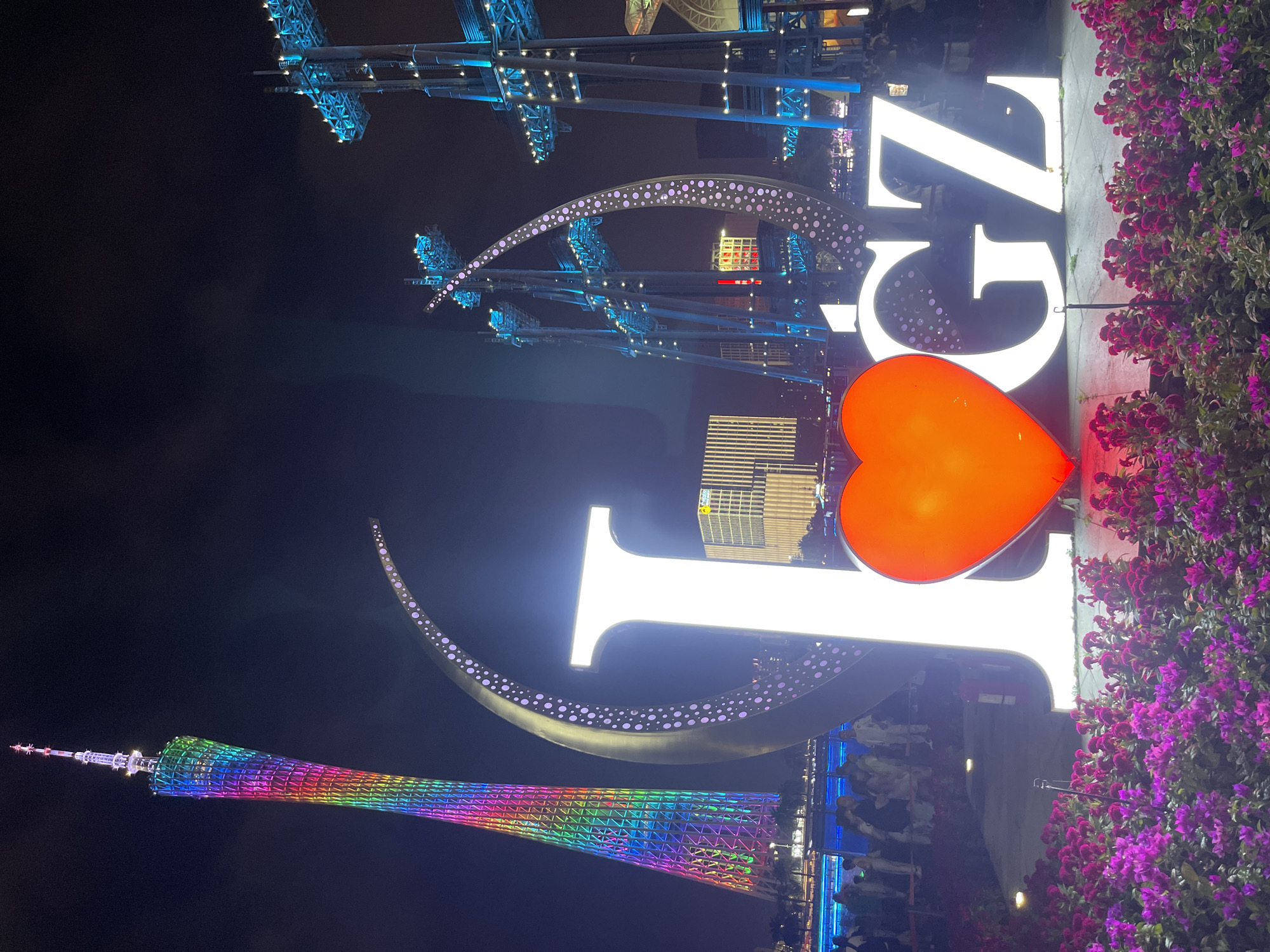 The image shows a nighttime scene in Tianhe District, China, featuring a prominent illuminated sculpture that spells out "I ❤️ GZ". The sculpture consists of large, bright white letters "I" and "GZ", with a large, red, illuminated heart in place of the word "love". The sculpture is surrounded by a bed of vibrant purple flowers in the foreground.
In the background, a tall, slender tower, likely the Canton Tower, is illuminated with a spectrum of rainbow colors. To the right of the "I ❤️ GZ" sculpture, there are several large, modern steel structures, also illuminated with bright blue lights. The scene appears to be a public space, possibly a waterfront promenade, with a body of water visible behind the sculpture and a crowd of people gathered. The buildings in the distance are also illuminated, contributing to the bright, festive atmosphere of the night.