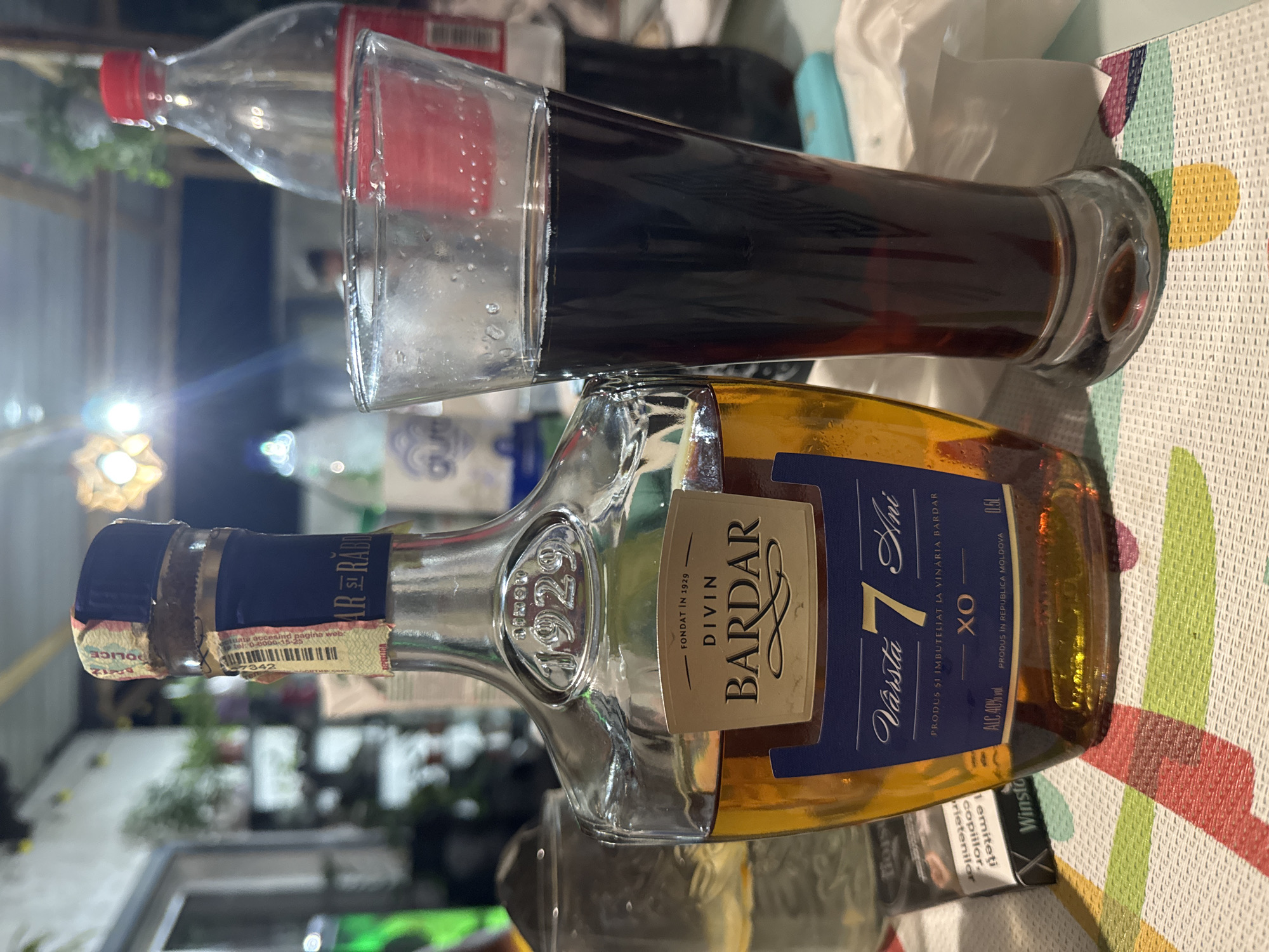 The image shows a close-up of a bottle of Divin Bardar XO brandy and a glass of dark liquid, likely cola, on a table. The bottle is made of clear glass with a gold label that reads "DIVIN BARDAR", "Vârsta 7 Ani", and "XO". The liquid inside is amber-colored. The glass is tall and filled with a dark brown beverage, topped with a thin layer of foam. In the background, a red plastic bottle of Coca-Cola is visible, along with other blurry elements of an indoor setting. A pack of Winston cigarettes is also present on the left side of the frame, partially obscured by the brandy bottle. The overall setting appears to be casual, possibly a home or a small establishment, with a tablecloth featuring a colorful geometric pattern. There is no indication of the city of Moscovei, Moldova within the image content itself.