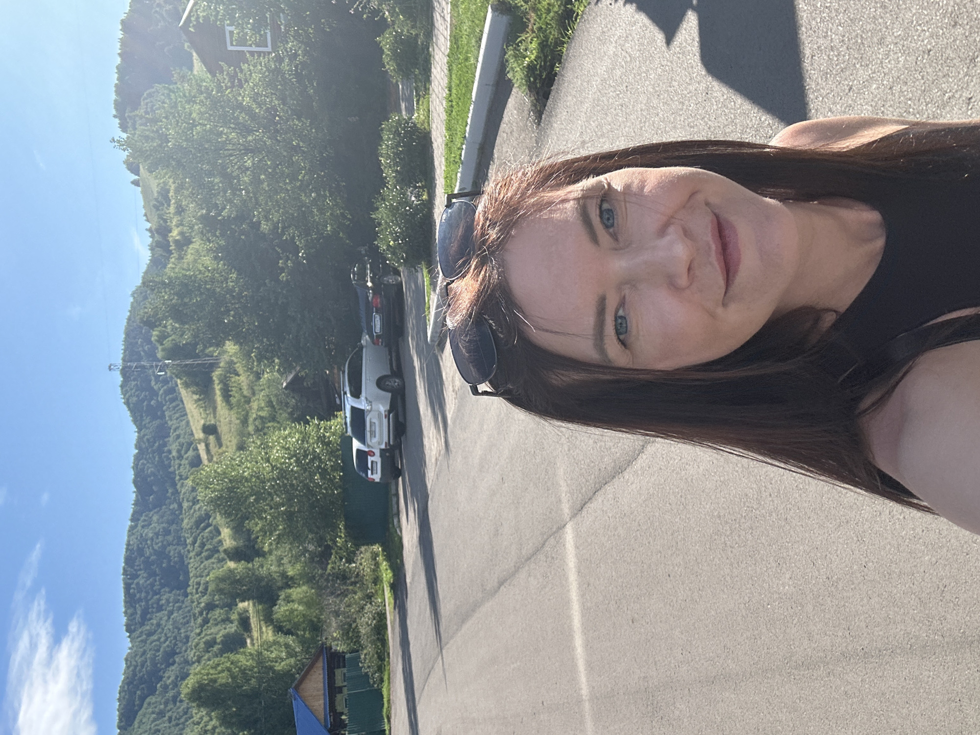 A selfie shows a woman with long brown hair and blue eyes, wearing a black top and sunglasses perched on her head. She is smiling at the camera. The background reveals a paved road with parked cars, including two white SUVs and a dark sedan. Beyond the road, there are trees, a wooden house with a blue roof, a green fence, and rolling hills covered in dense green forest. The sky is bright blue with scattered white clouds, and the sun casts shadows on the road. The location is identified as Danilovka, Altai Krai, Russia.