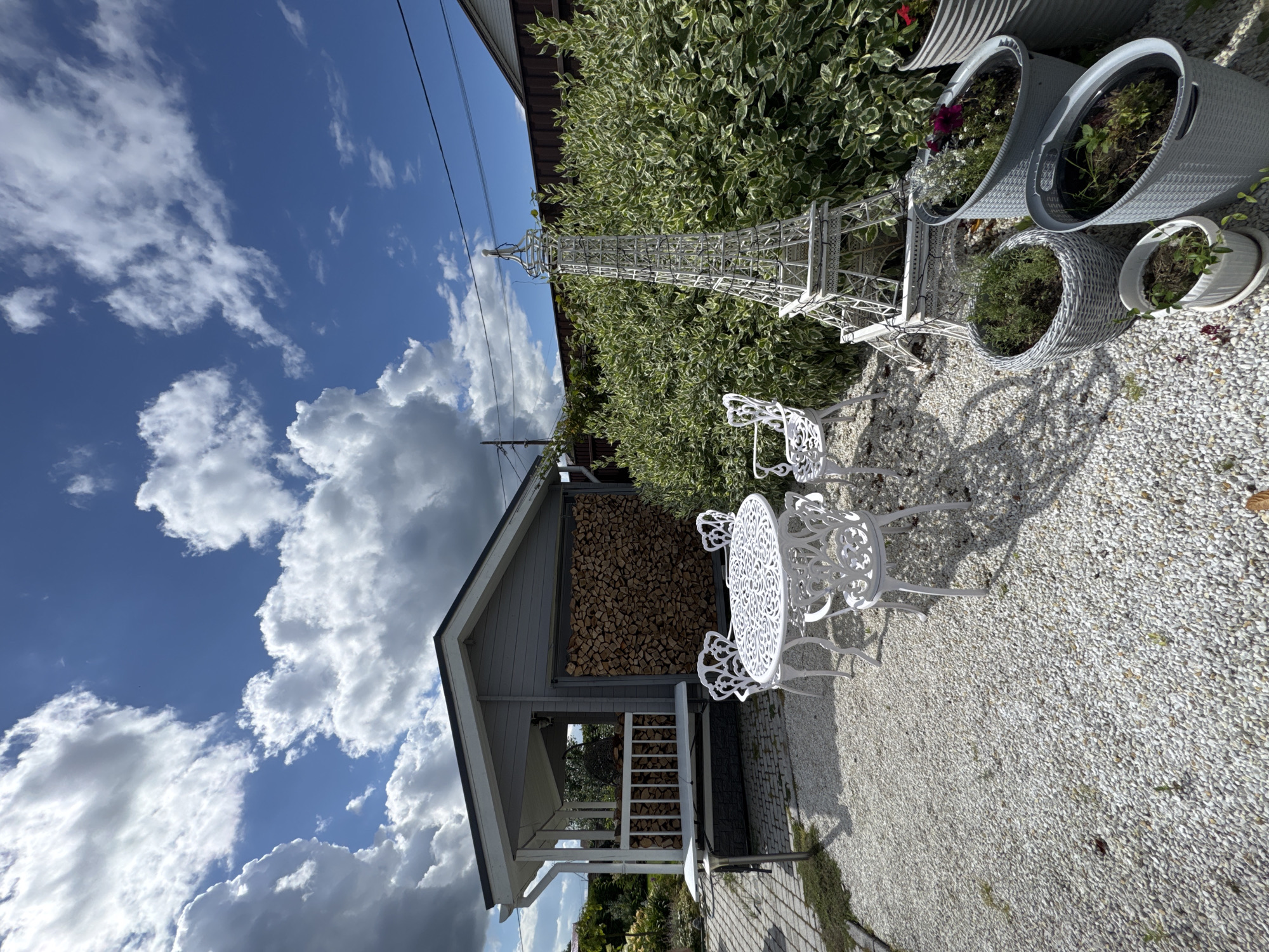 An outdoor scene in Oliki, Russia, features a gravel-covered backyard with a small gray structure, a white wrought-iron table and chairs, and various potted plants. The structure, which appears to be a shed or a gazebo, is adorned with a railing and houses a large stack of firewood. Adjacent to the structure, a white wrought-iron table is set with four chairs, all decorated with intricate patterns. To the right, a miniature replica of the Eiffel Tower stands prominently, surrounded by several gray planters containing greenery and flowers. The ground is covered in small white pebbles, with patches of grass and a few fallen leaves. The sky above is a clear blue, dotted with fluffy white clouds, indicating fair weather. The overall ambiance is that of a well-maintained, charming garden space.