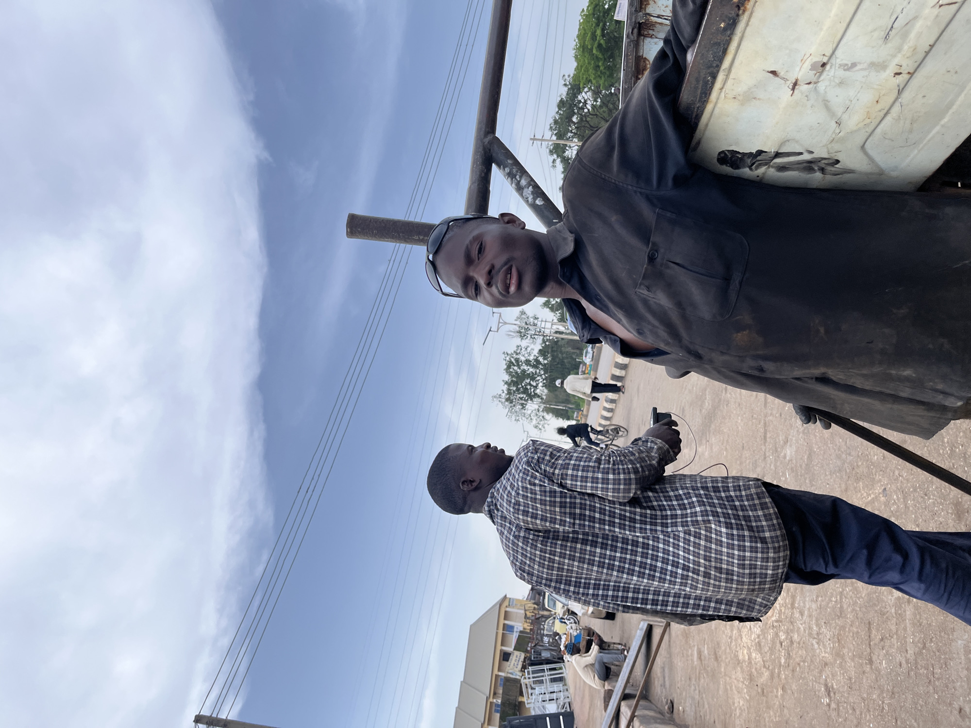 The media file shows two men outdoors in what appears to be a dusty or industrial setting, possibly a scrapyard or market. The sky is visible with scattered clouds, suggesting daytime. The first man, in the foreground, is wearing a black long-sleeved shirt and dark pants. He has sunglasses on his forehead and is looking towards the camera with a neutral expression. The second man is in the background, wearing a plaid shirt and dark pants. He is looking to the side and holding a black object, possibly a phone or camera, in his hand.
Behind the men, there are metal structures, possibly parts of vehicles or discarded machinery, and some vegetation in the distance. The overall impression is that of a working environment, likely in a location with limited infrastructure, given the visible power lines and the general condition of the metal objects. The lighting and the appearance of the sky suggest it is likely midday or afternoon. No visible text is present in the image. The scene appears to be taking place in Gura Top, Nigeria.
