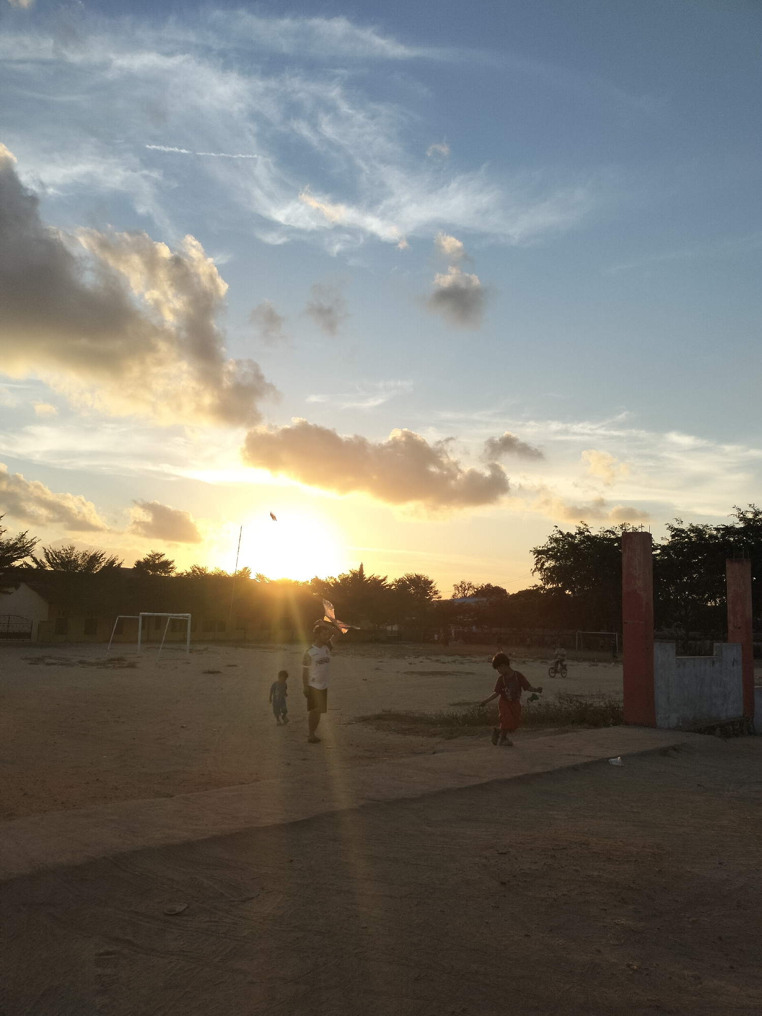 The image depicts an outdoor scene at what appears to be a field or playground in Karang Indah, Indonesia, during sunset. The sky is a gradient of blue with scattered clouds, some illuminated by the setting sun. The sun itself is a bright, intense light source, casting long shadows and lens flares across the scene.
In the foreground, a dirt and concrete area is visible. Several children are present. One child, wearing a red shirt and shorts, is running or walking towards the right side of the frame. Another child, wearing a blue outfit, is walking towards the left. An adult or older child, wearing a white t-shirt with dark lettering and dark shorts, is standing near the center, holding a kite. The kite is visible in the air.
In the middle ground, there is a soccer goal with a white frame. Beyond the goal, there are trees and a building with a light-colored facade. In the background, more trees are visible against the sky. To the far right of the image, there is a structure with red-painted pillars and a grey concrete section. The overall impression is one of a casual, late afternoon activity in a somewhat rural or semi-urban setting within Karang Indah.