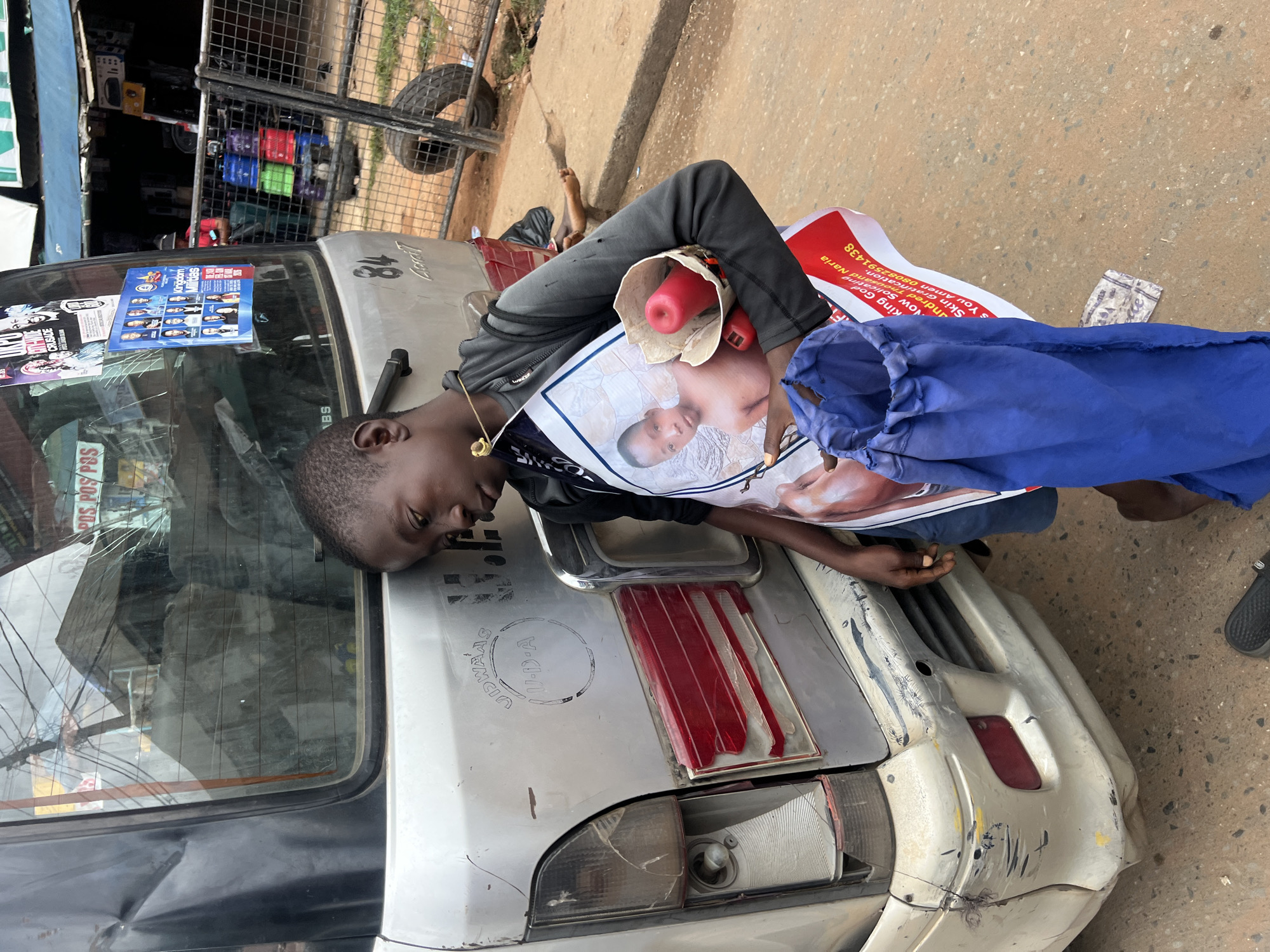 The image captures a young boy leaning against the rear of a dented, silver van in Nkpor, Nigeria. He appears tired, with his eyes closed and head resting on the vehicle. The boy is holding a poster with a picture of a young boy on it, and a red megaphone is tucked into a white paper cone. A blue drawstring bag hangs from his arm. The van's taillights are broken and the bumper is heavily scratched, indicative of its condition and the environment. In the background, a street market is visible with stalls, various items for sale, and other people. The ground is paved with concrete and dirt.