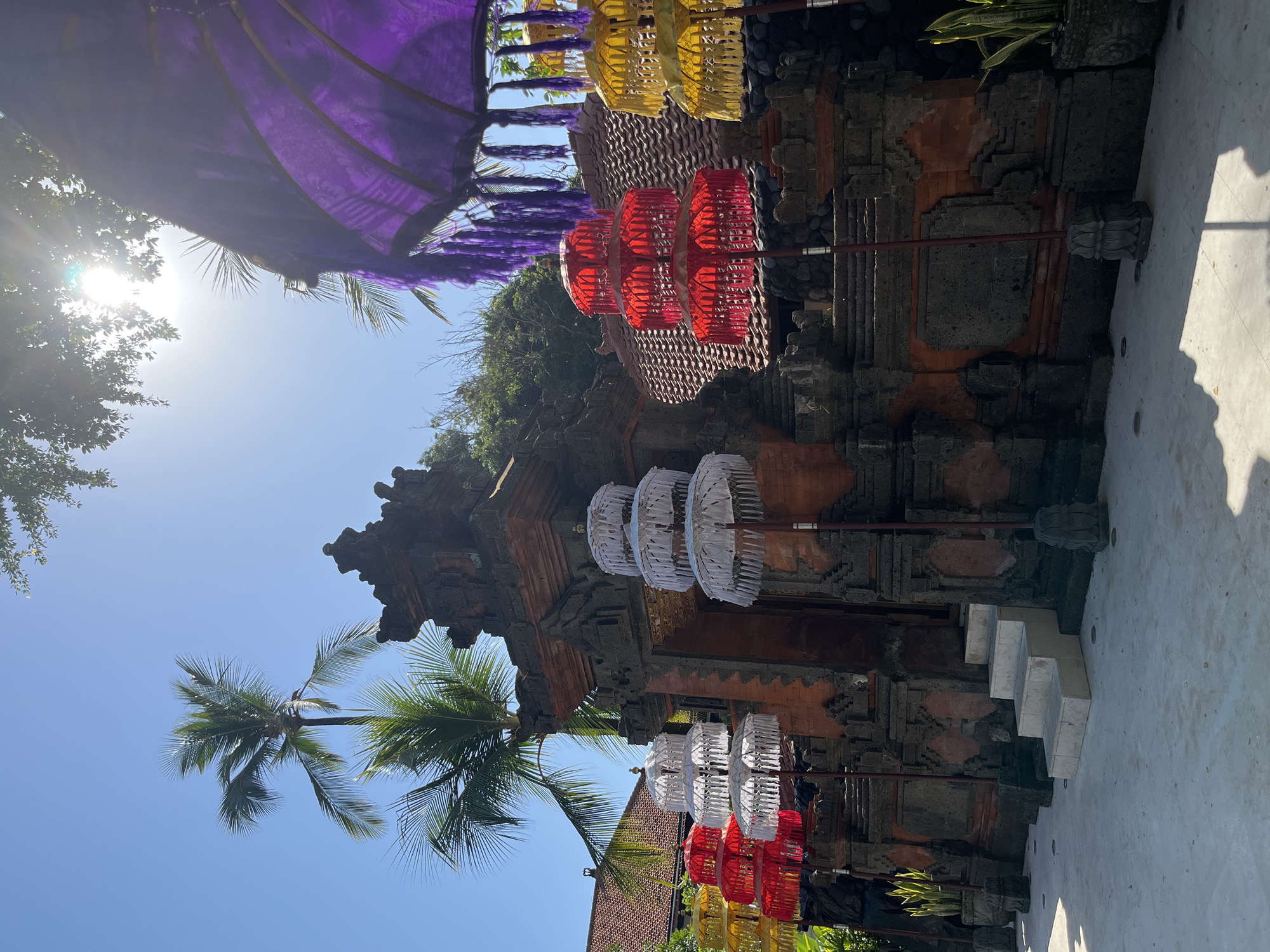 In Kuta Selatan, Indonesia, a traditional Balinese gate stands adorned with colorful tiered umbrellas. The ornate brick gate, likely an entrance to a temple or significant building, is decorated with intricate stone carvings. In front of the gate, several white, red, yellow, and purple tiered umbrellas are displayed on tall poles, adding a festive and ceremonial atmosphere. These umbrellas are a common sight in Bali, often used for religious ceremonies and cultural events. Palm trees are visible behind the gate, and the bright blue sky suggests a sunny day. The foreground shows a paved courtyard. The scene evokes a sense of cultural richness and vibrant celebration.